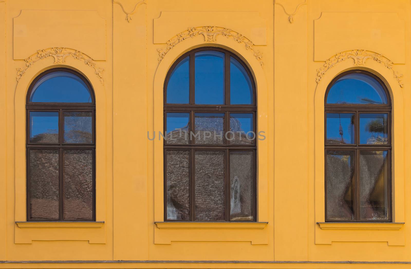 Ornate yellow facade of an old building with three windows with archs, reflecting a blue sky and a roof a the oposite building.