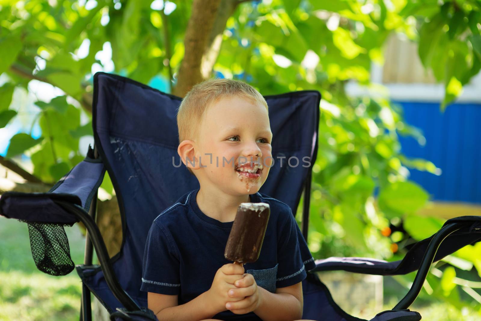 Adorable little boy eating ice cream pops in home's garden, outdoors. Happy blond kid eating chocolate ice cream bar