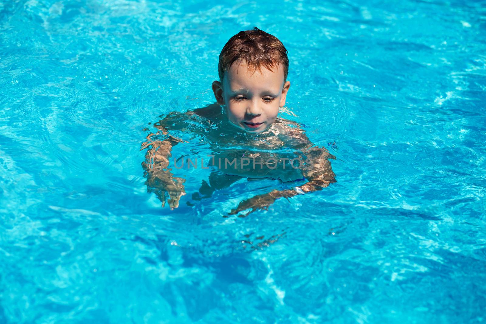 Happy kid playing in blue water of swimming pool on a tropical resort at the sea. Summer vacations concept. Cute boy swimming in pool water. Child splashing and having fun in swimming pool