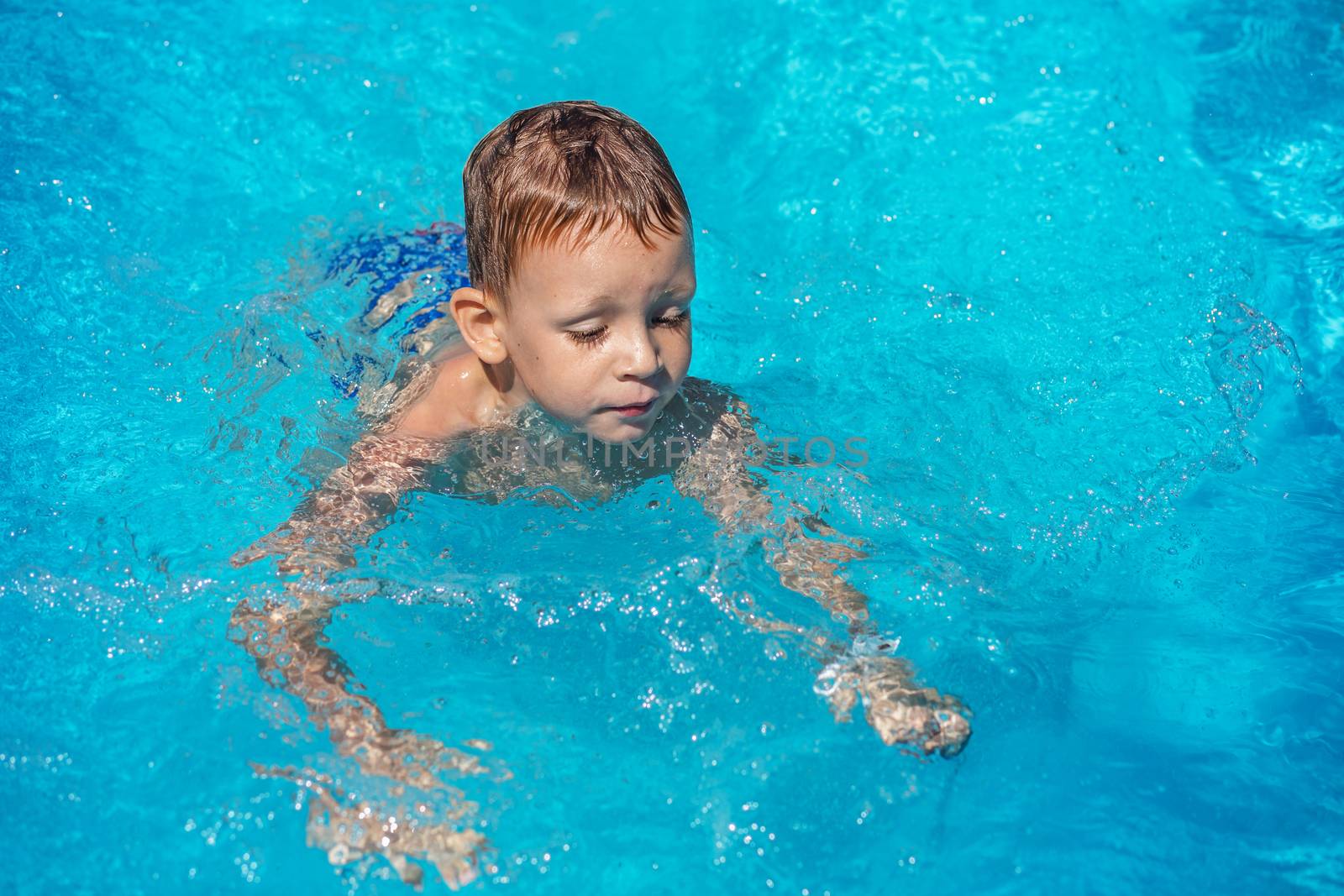Happy kid playing in blue water of swimming pool. Little boy learning to swim. Summer vacations concept. Cute boy swimming in pool water. Child splashing and having fun in swimming pool