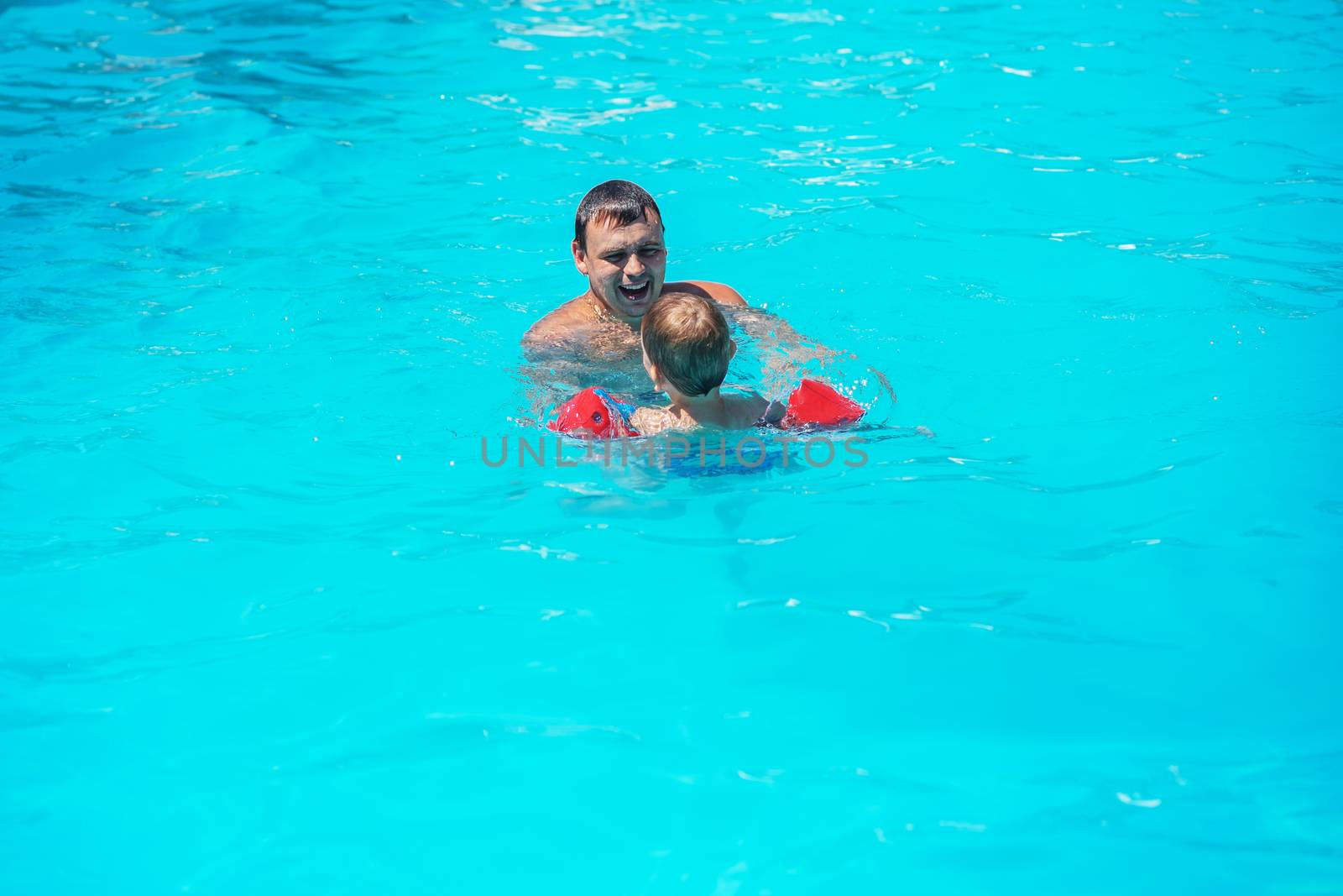 Dad and son swimming together in the pool. Happy family playing in blue water of swimming pool. Kid learning to swim. Summer vacations concept. Dad and son having fun in swim pool