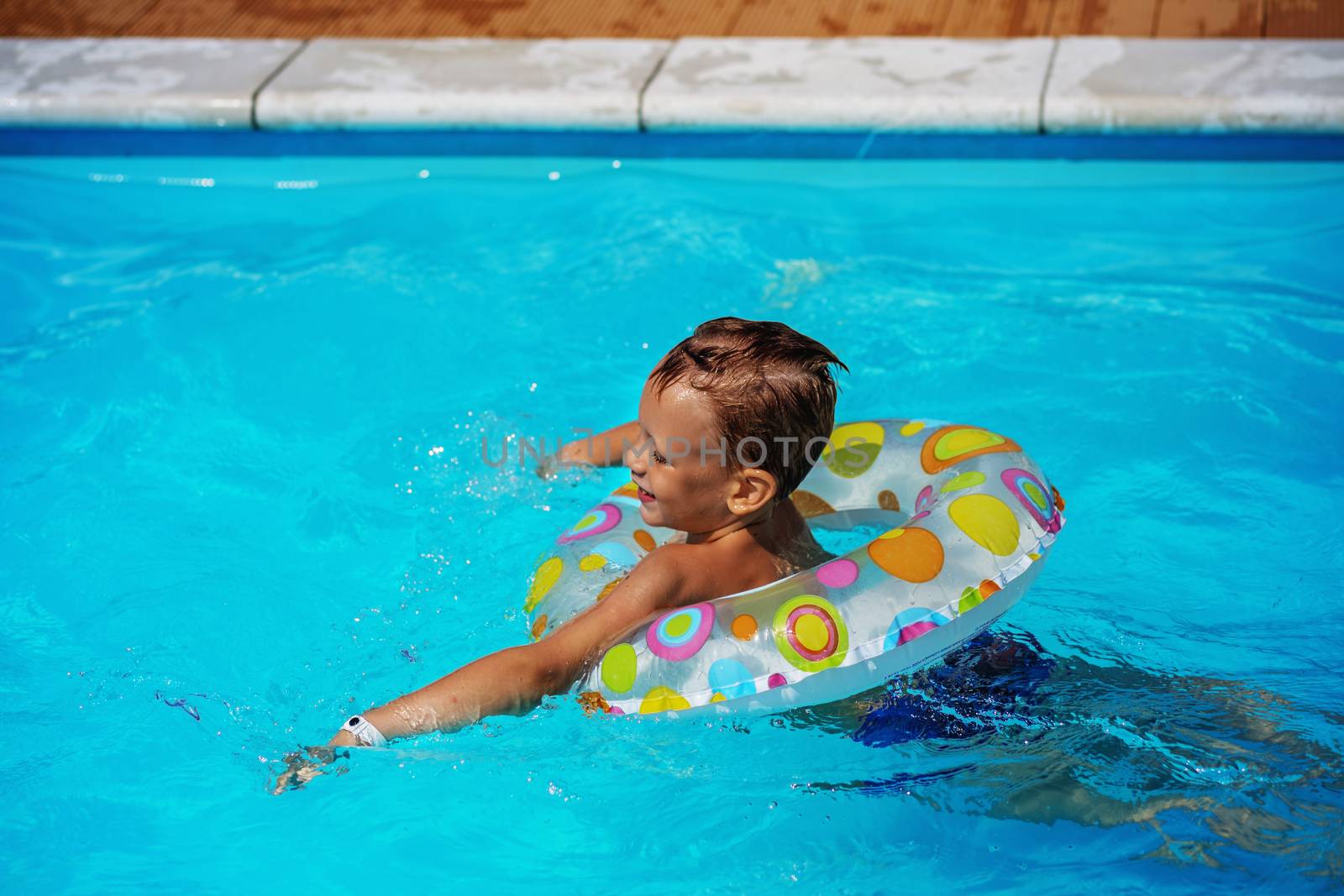 Happy kid playing in blue water of swimming pool with swim ring. Little boy learning to swim. Summer vacations concept. Cute boy swimming in pool water. Child splashing and having fun in swimming pool