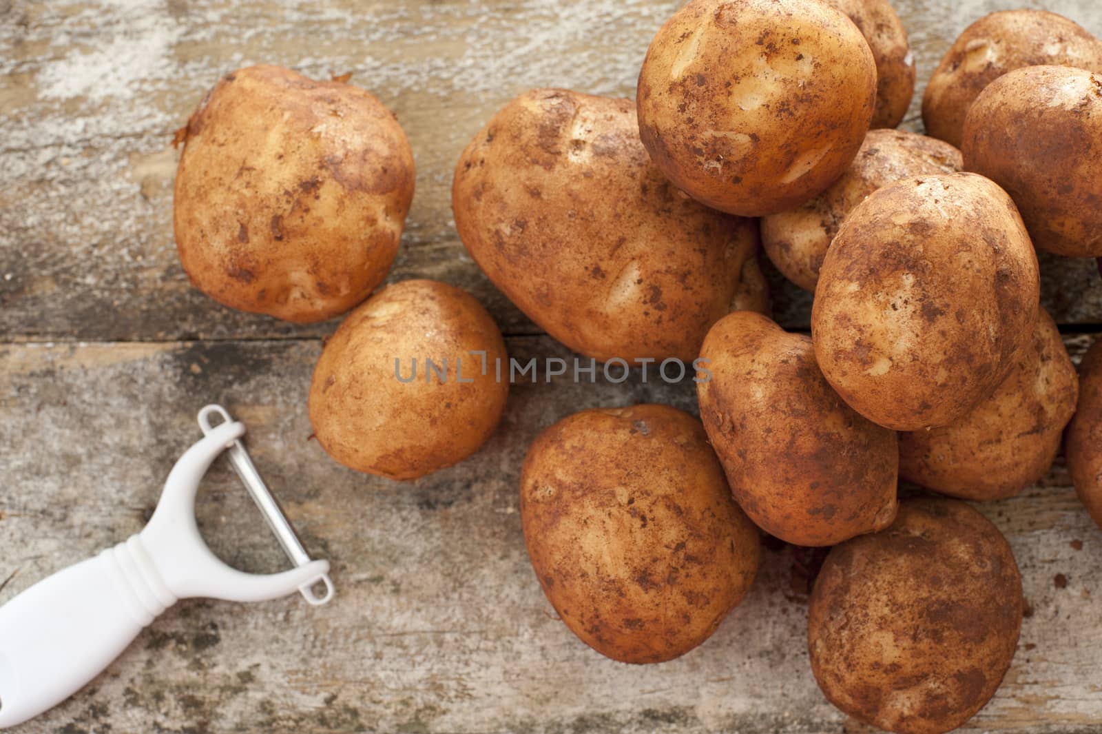 Pile of round baking potatoes beside peeler against a rustic background as seen from an overhead view