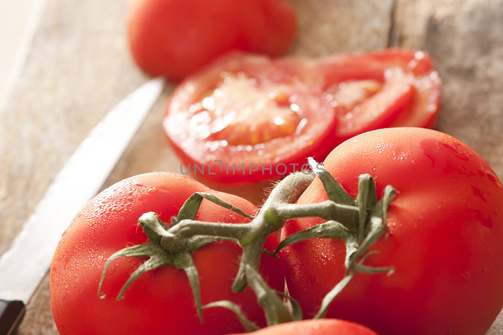 Close up view of succulent red tomatoes on vine beside sliced one and shiny sharp knife