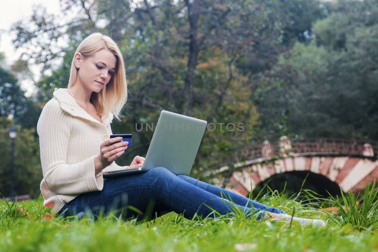 Girl sitting in park and holding credit card makes online shopping using wireless laptop.