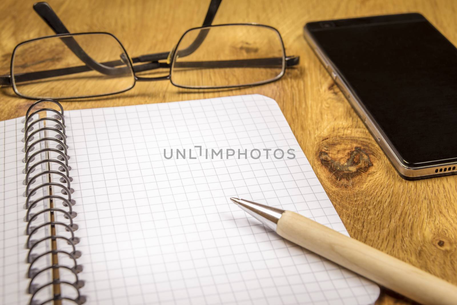 Close-up image with an opened graph, spiral notebook, eyeglasses, pen and phone on a vintage, wooden table. Perfect as a frame or background.