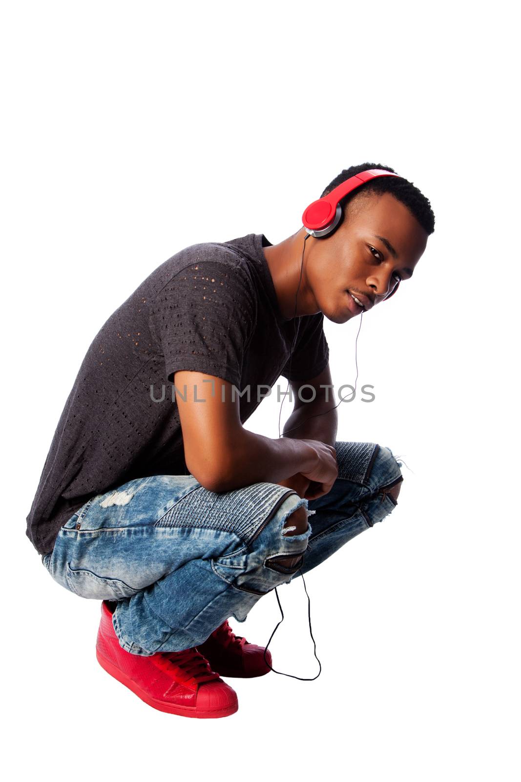 Handsome African teenager listening to music wearing red headphones while squatting, on white.