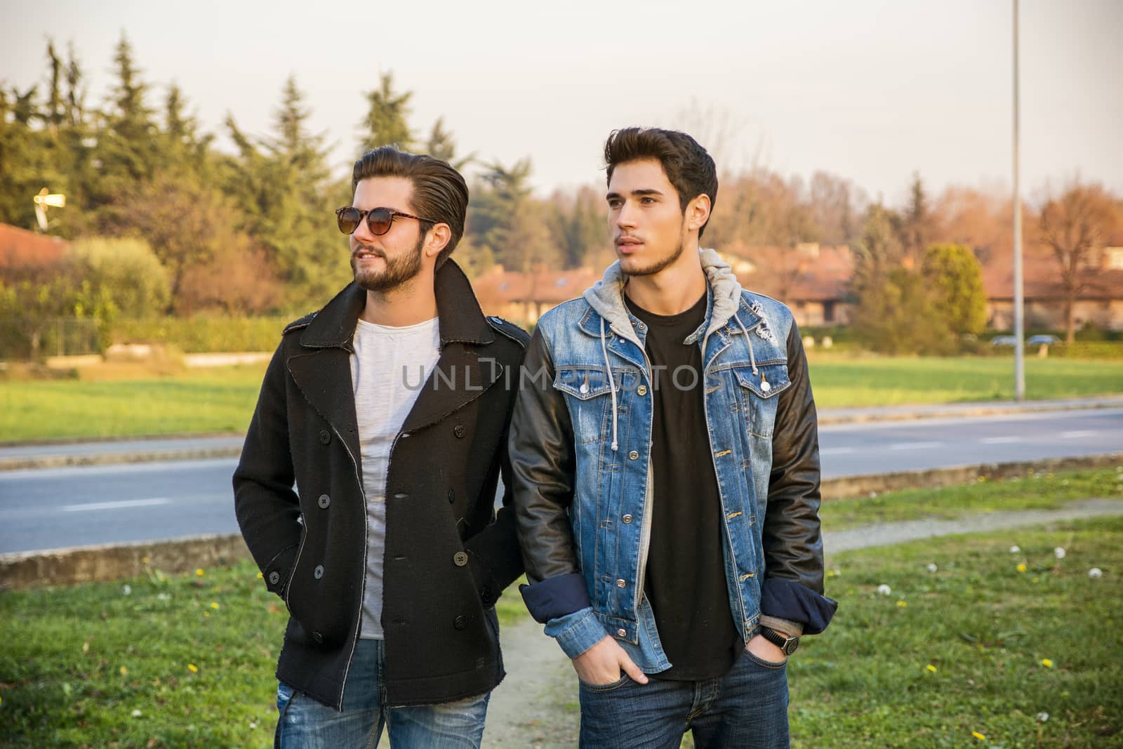 Two handsome casual trendy young men, 2 friends, in an urban park walking and chatting together