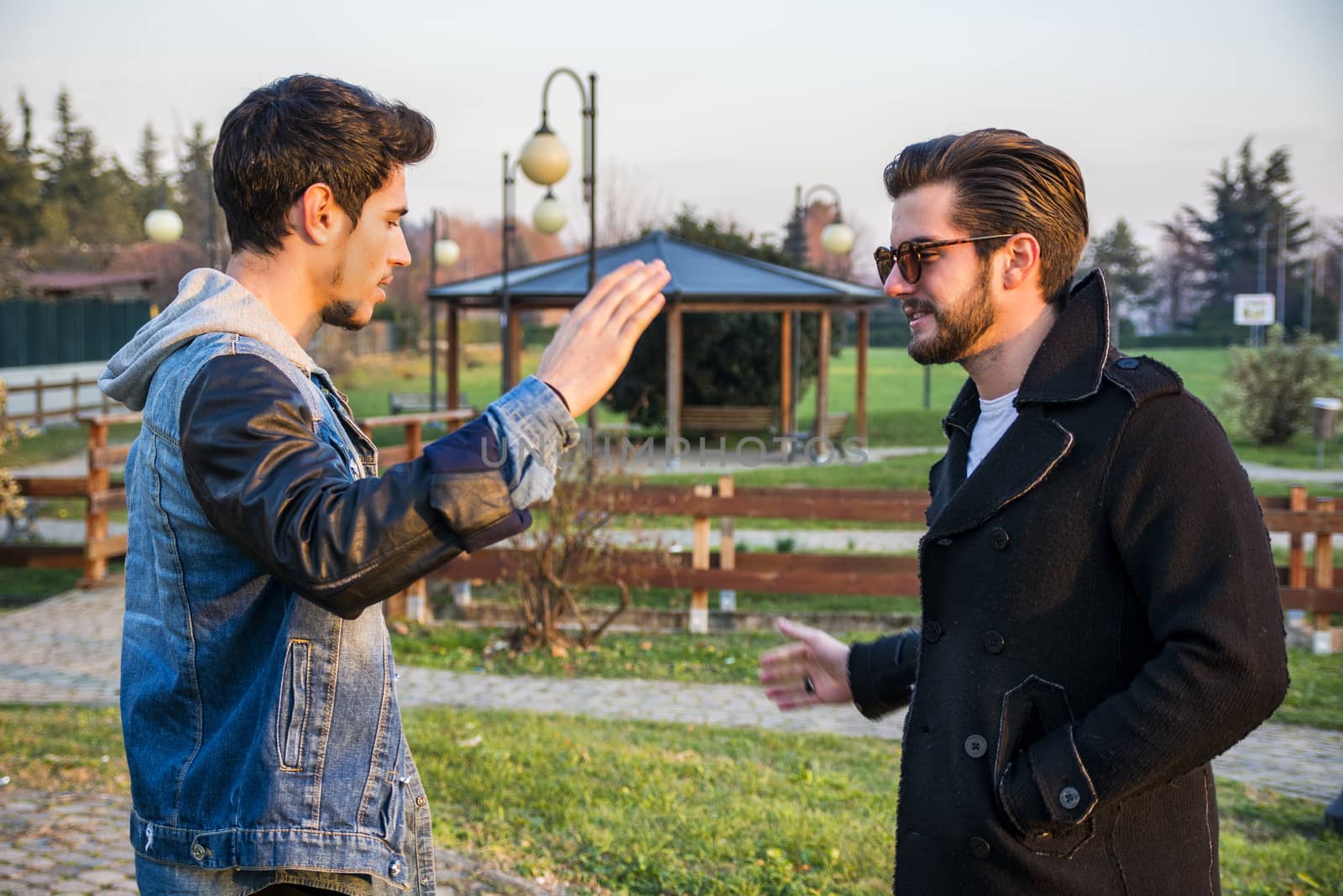 Two handsome casual trendy young men greeting outdoors in an urban park gripping hands with happy welcoming smiles