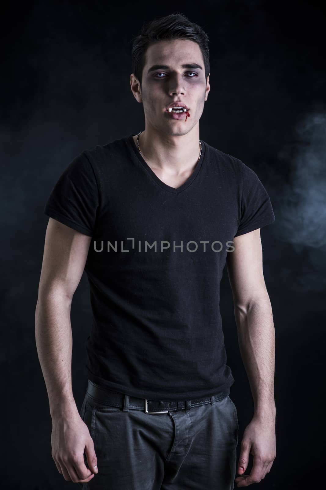 Portrait of a Young Vampire Man with Black T-Shirt, Looking at the Camera, on a Dark Smoky Background.