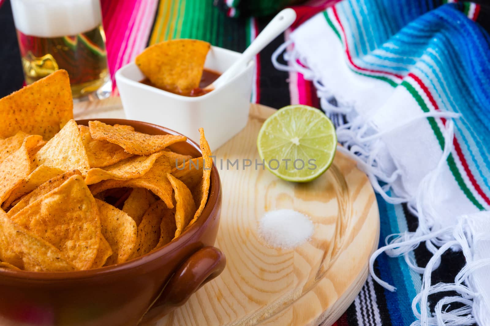Nachos chips in a bowl over a chopping board with tomato sauce beer lime and salt with poncho background