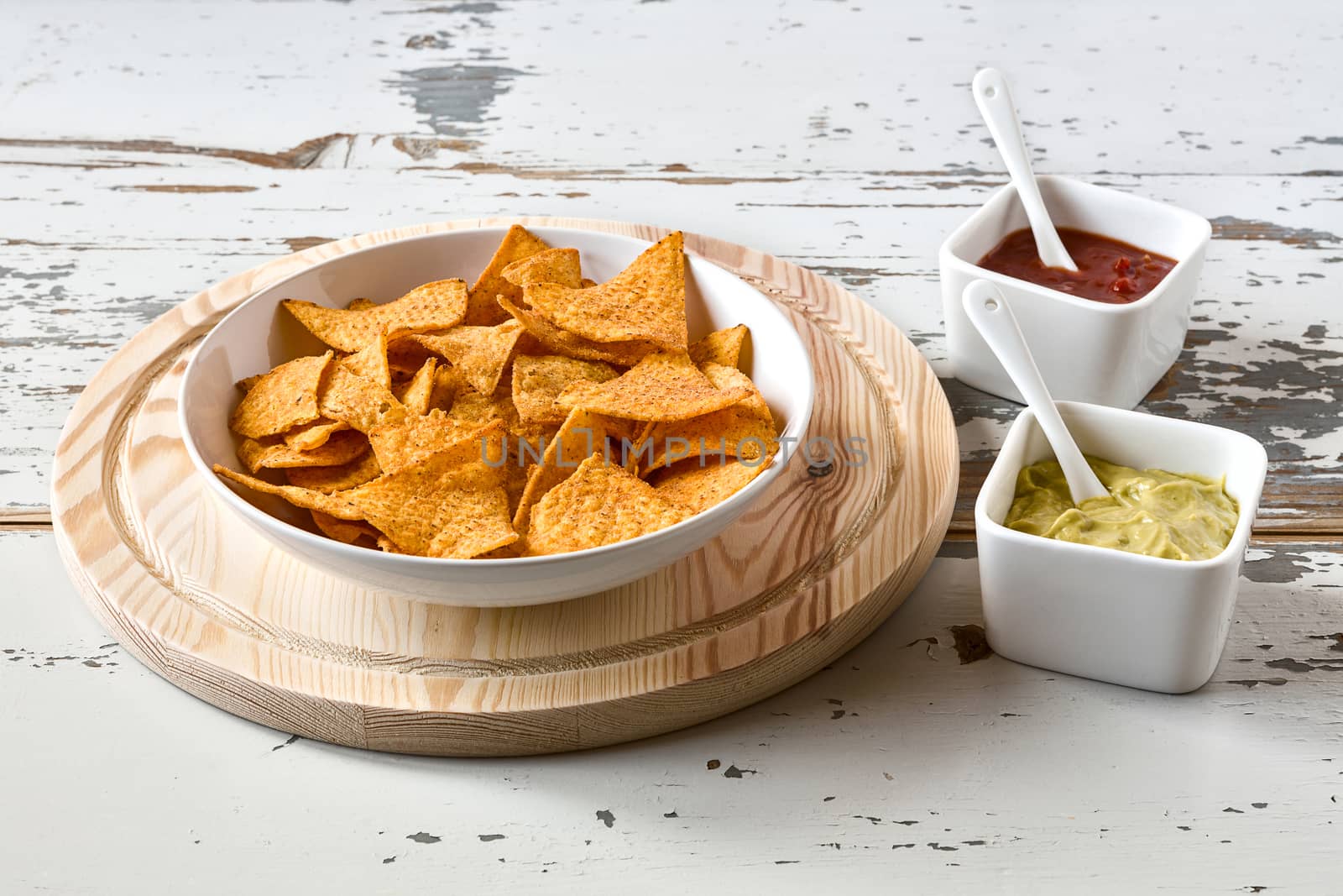 Nachos chips in a white bowl over a chopping board with sauces on a wood background