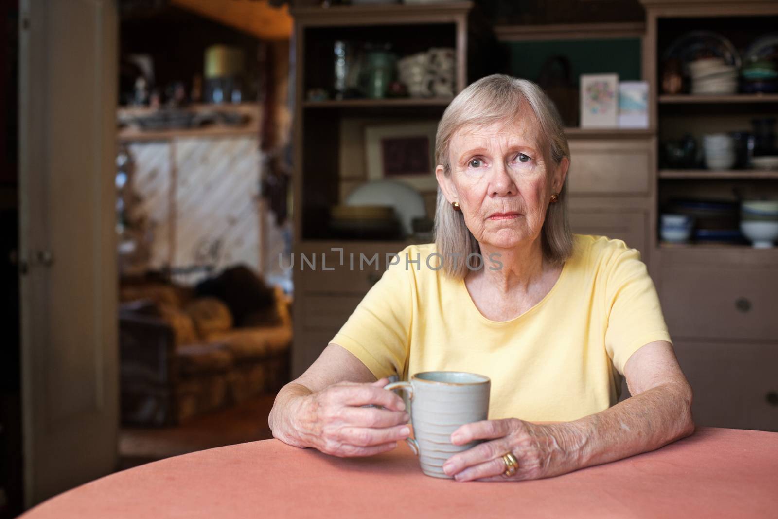 Senior woman in yellow shirt with depressed expression holding cup indoors