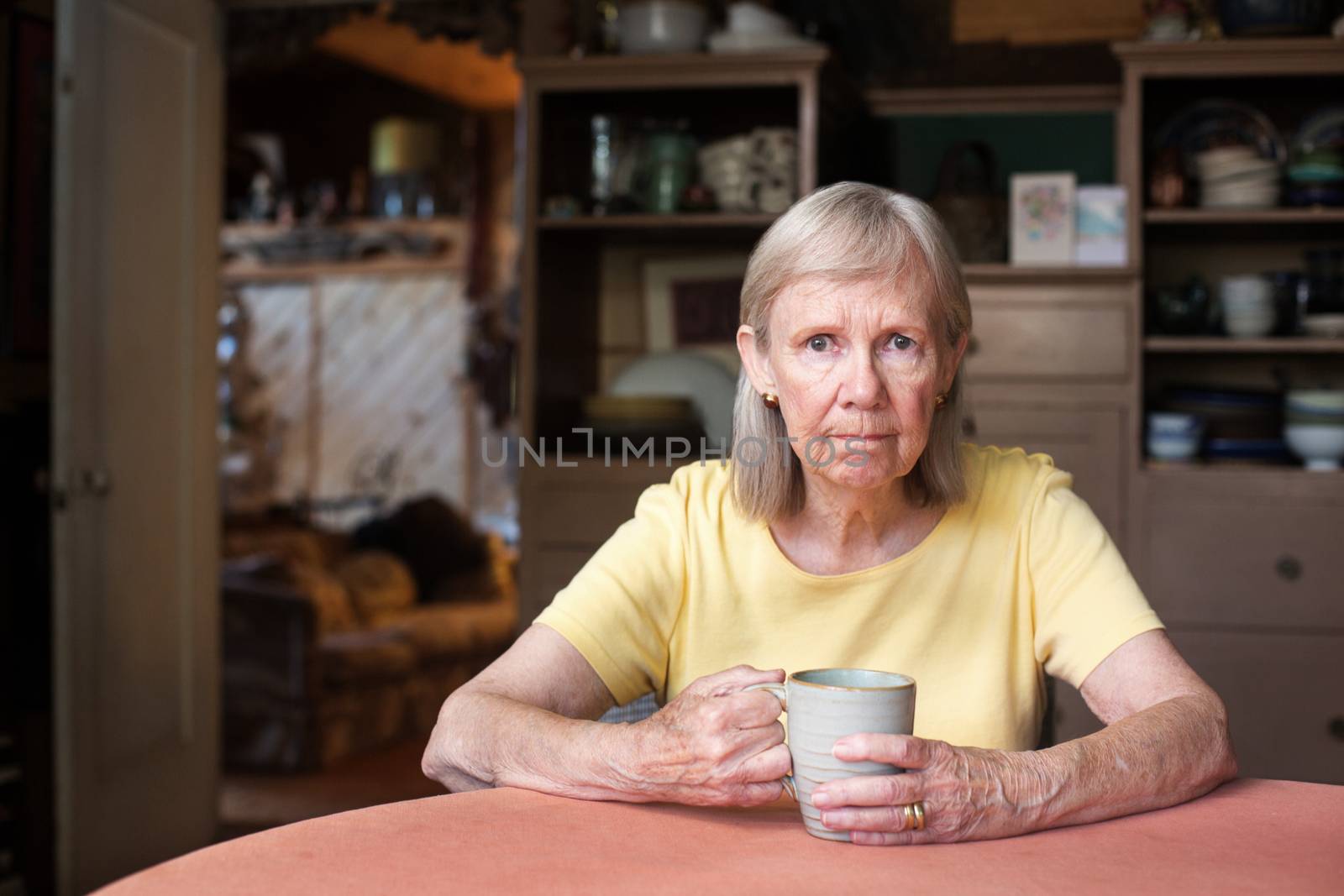 Frustrated single senior woman in yellow shirt holding cup while seated at table