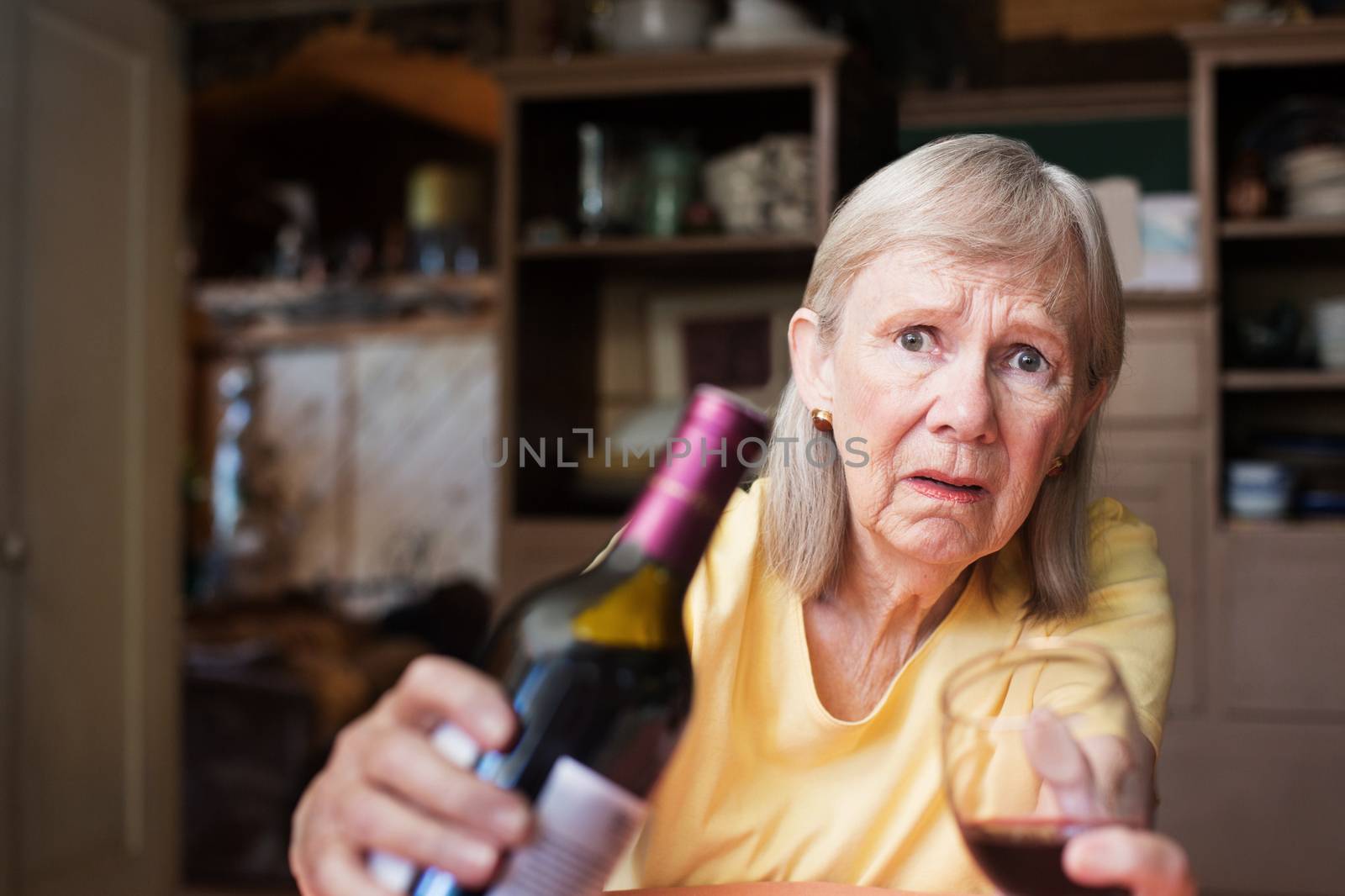 Worried mature adult female offering bottle of alcohol and glass sitting alone