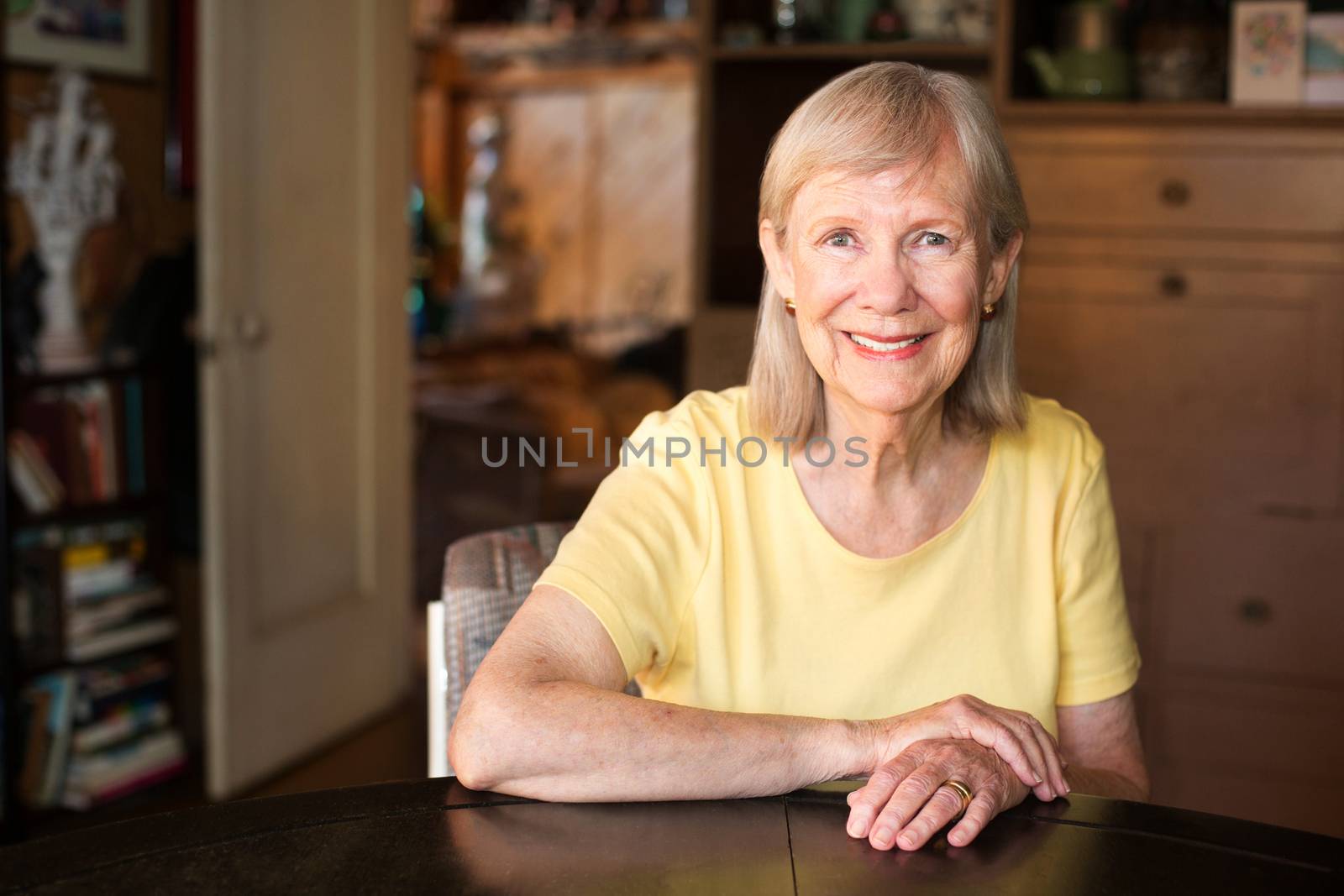 Smiling mature senior woman in yellow shirt seated at table indoors