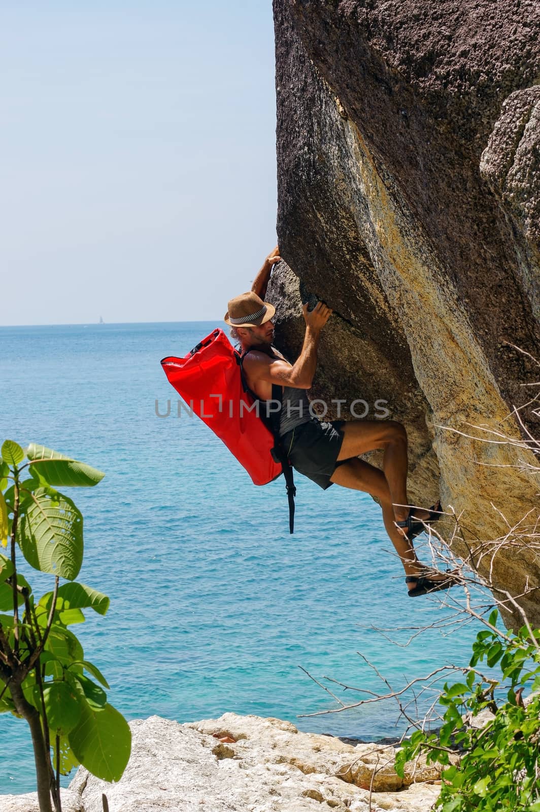 a strong and happy man climbing on high rock over the sea with a hut and a red seabag.