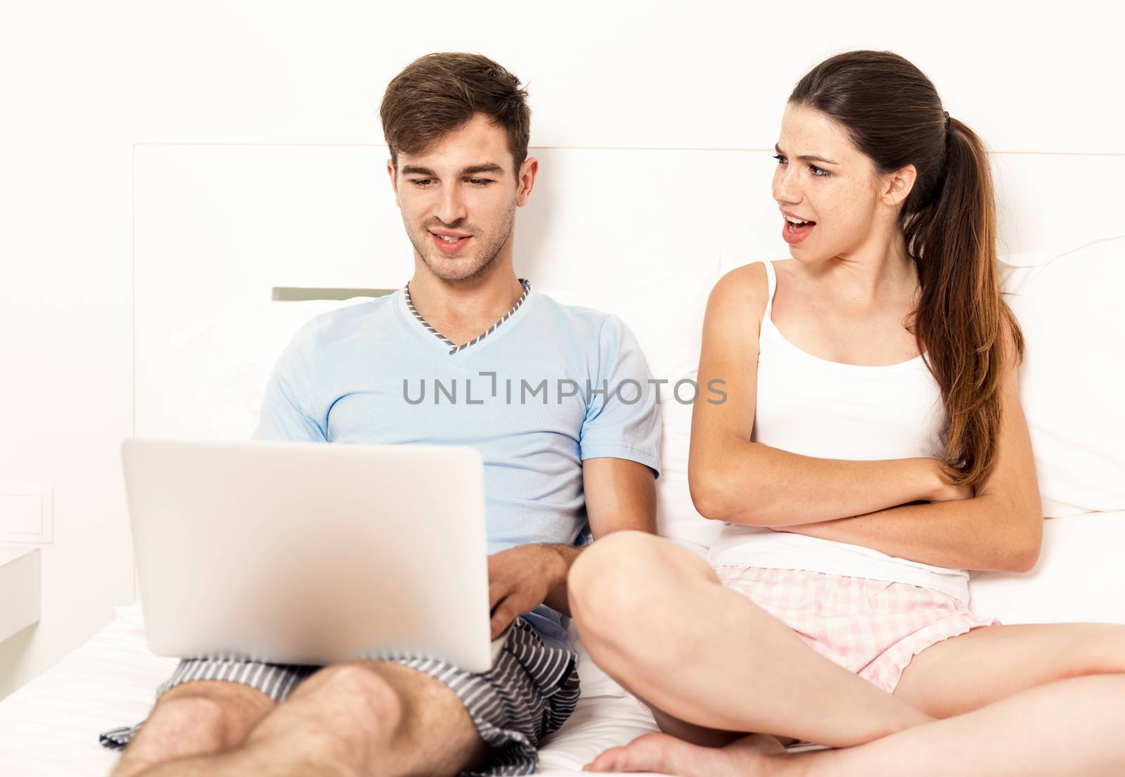 Addicted young man on bed with a laptop while the woman looks angry