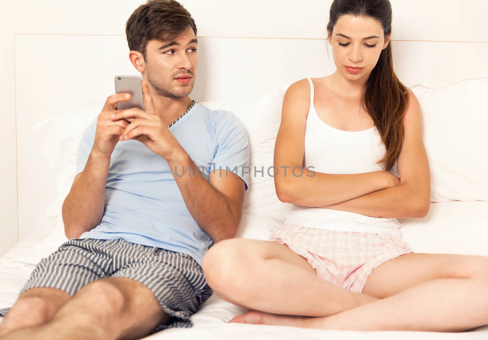 Addicted young man on bed with a cellphone while the woman looks angry