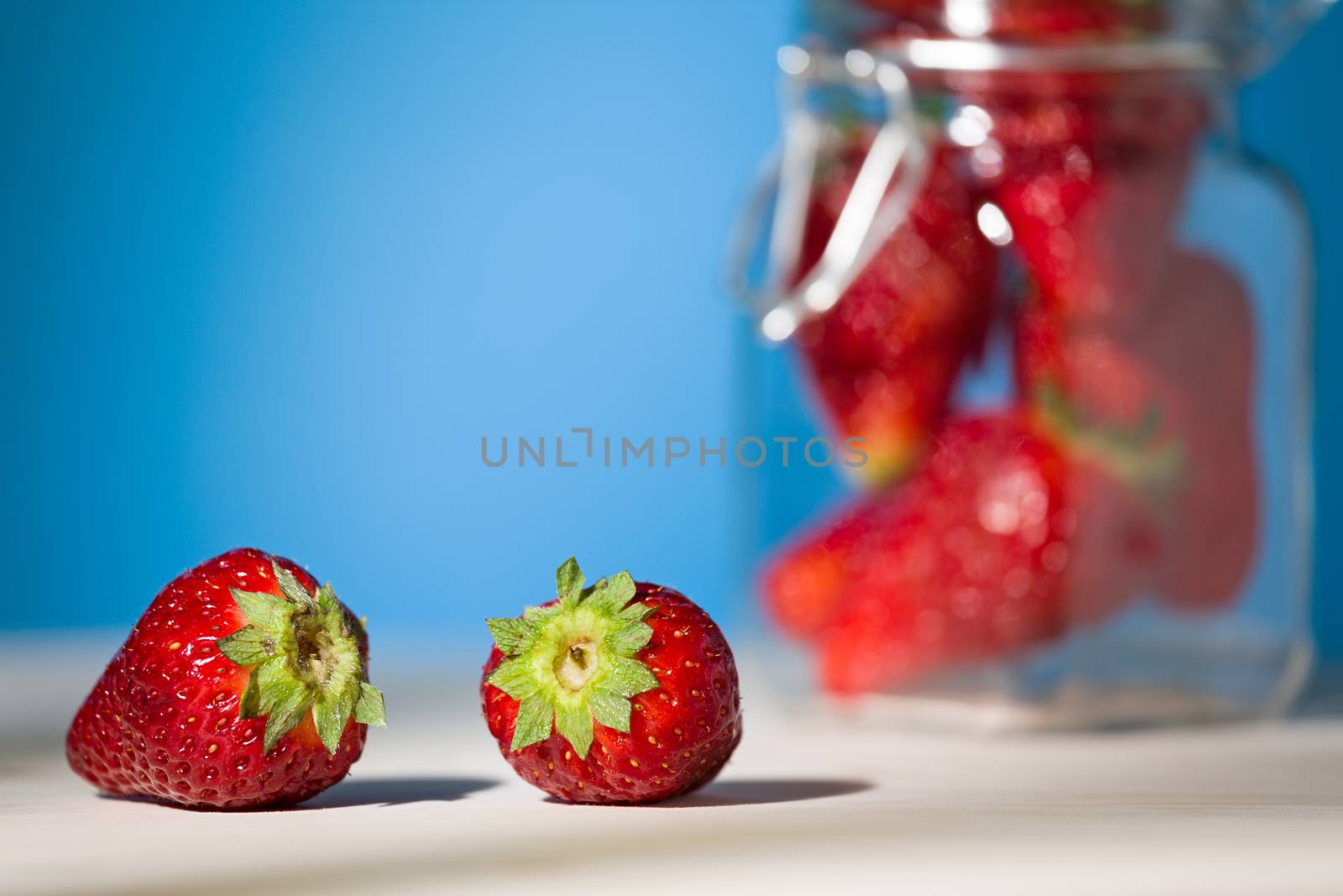 Close up of two strawberries on a table with blue background and a glass jar full of strawberries on the background