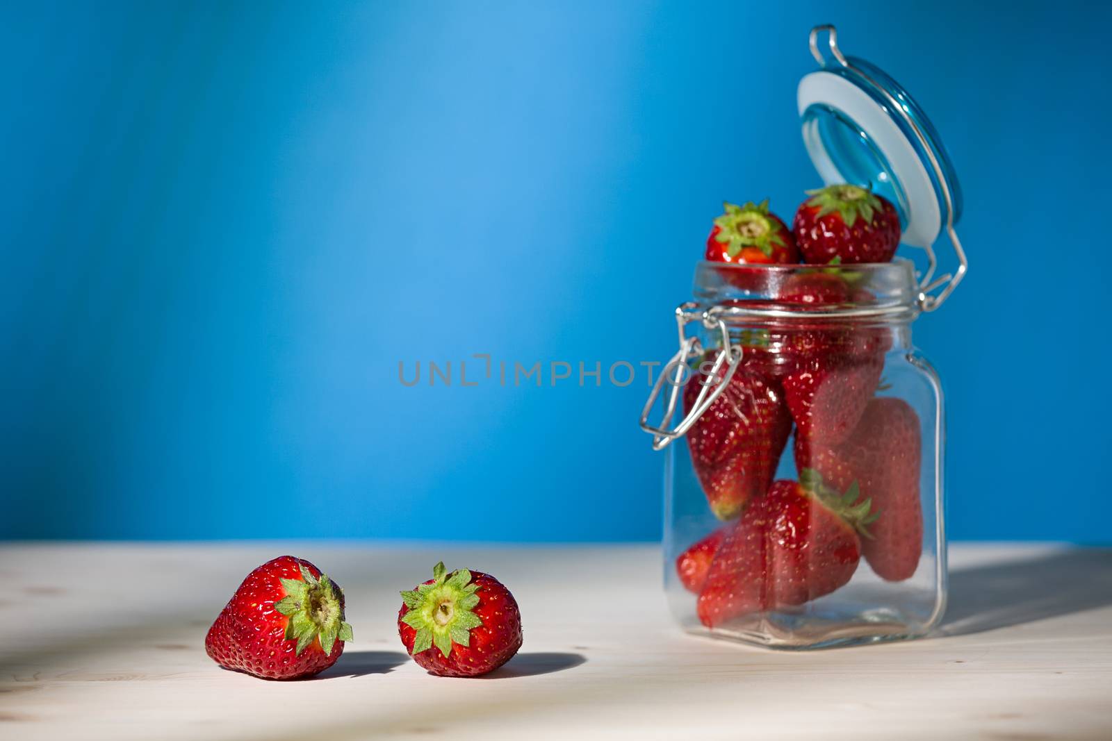 Strawberries on a table and a glass jar full of strawberries with blue background