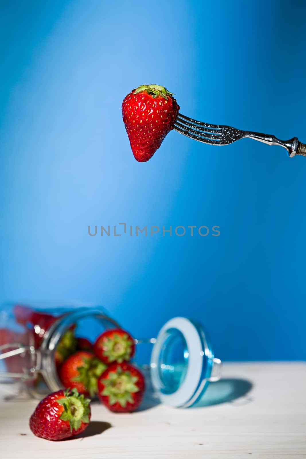 Strawberry suspended from a fork and under a jar of strawberries with blue background