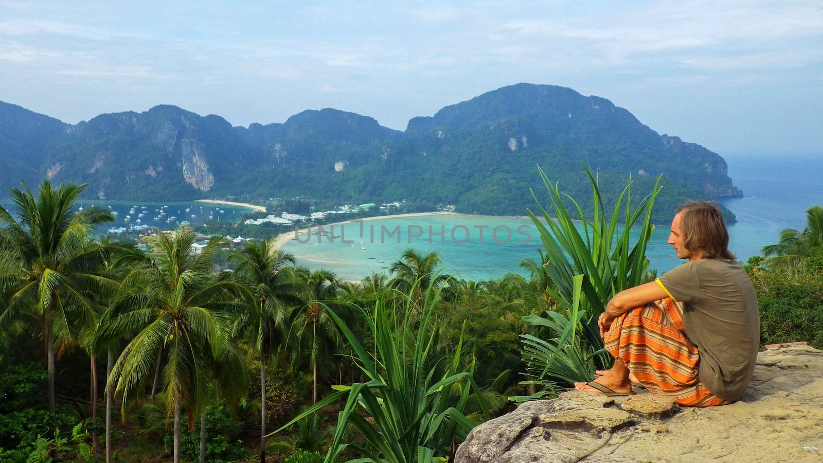 a young man sitting on a rock outdoors on Phi Phi Don Island, Thailand with a bay view from above.