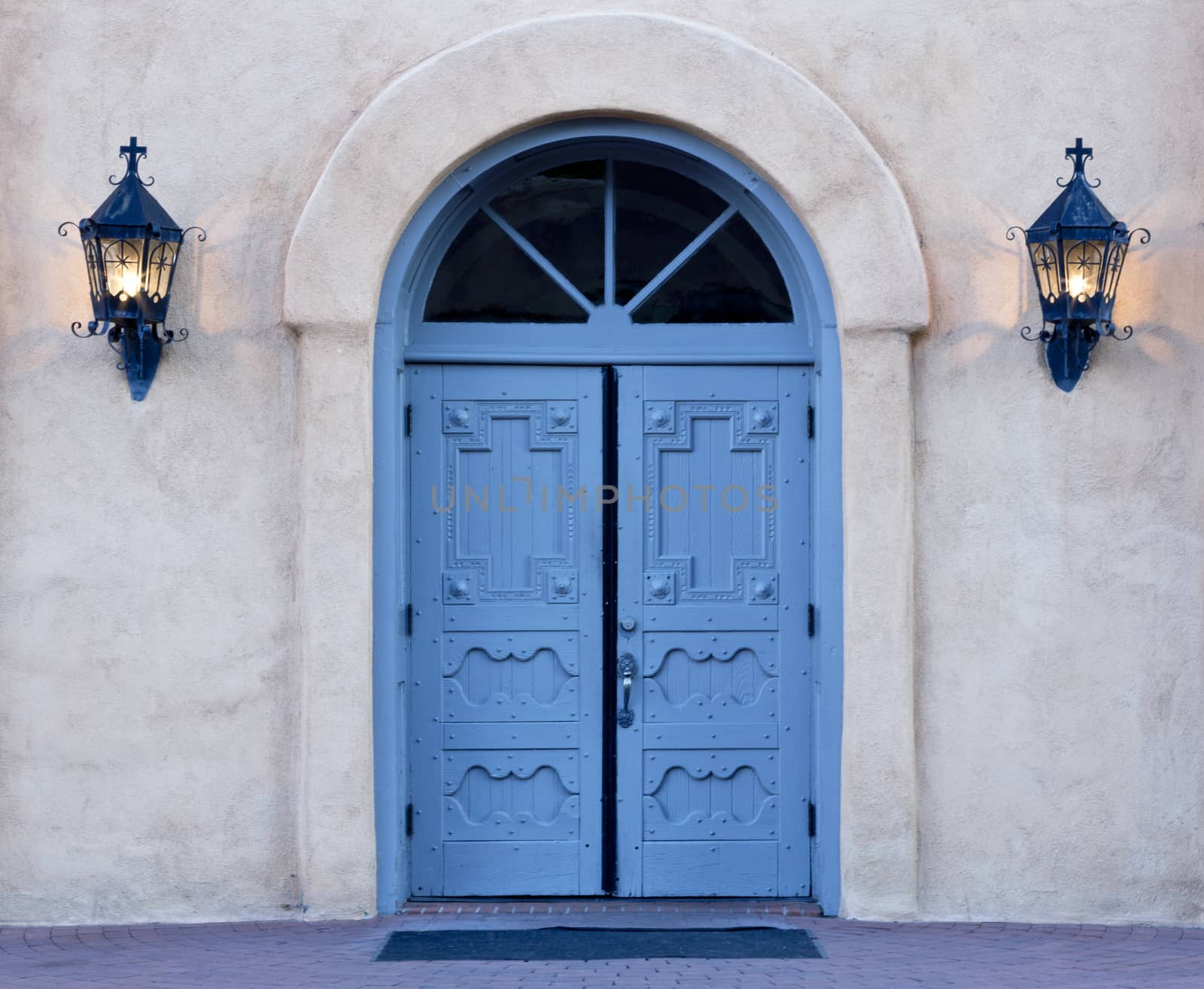 Morning rays begin to illuminate the azure blue, double doors of San Felipe de Neri church in Old Town, Albuquerque, New Mexico. Iron lanterns still glow on adobe wall. This historic church building is listed on National Register of Historic Places.  Preservation of Pueblo Spanish style architecture accents America’s Southwest and is a picturesque tourist attraction. Date is July 4, 2015.