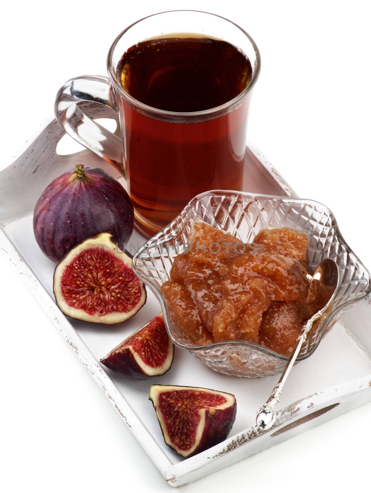 Arrangement of Delicious Homemade Fig Jam in Glass Kremanka with Fresh Fig Fruits and Glass Cup of Tea in White Wooden Tray closeup on White background
