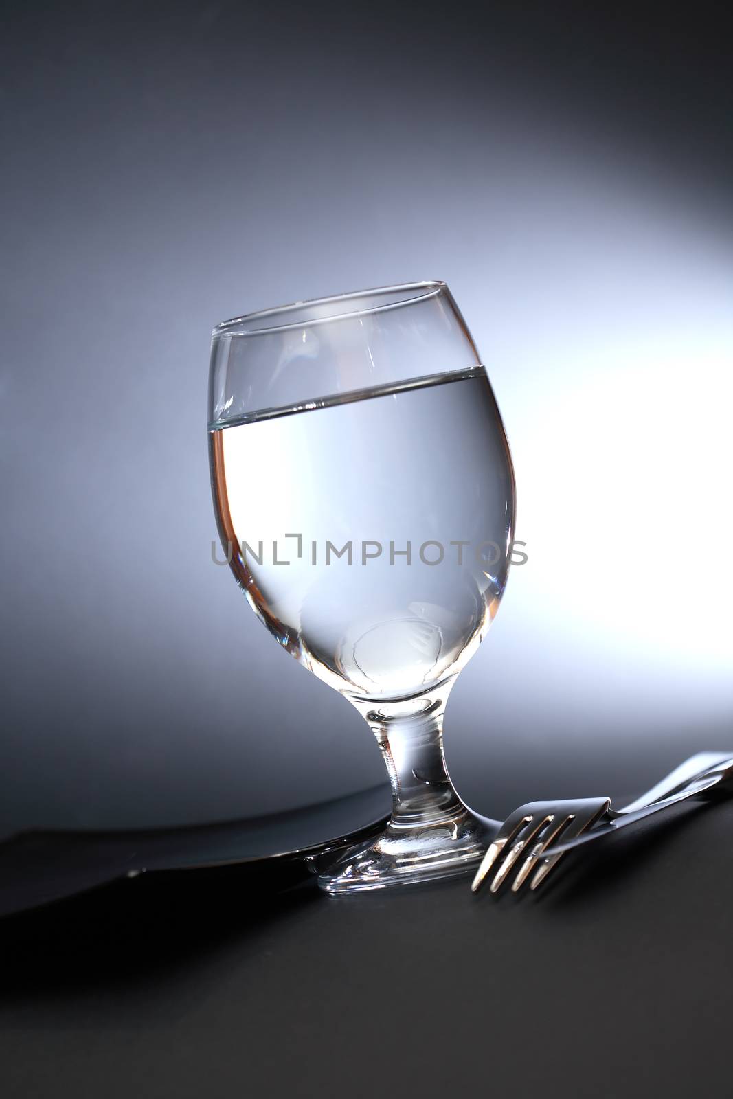 One glass of water near fork and knife on dark background