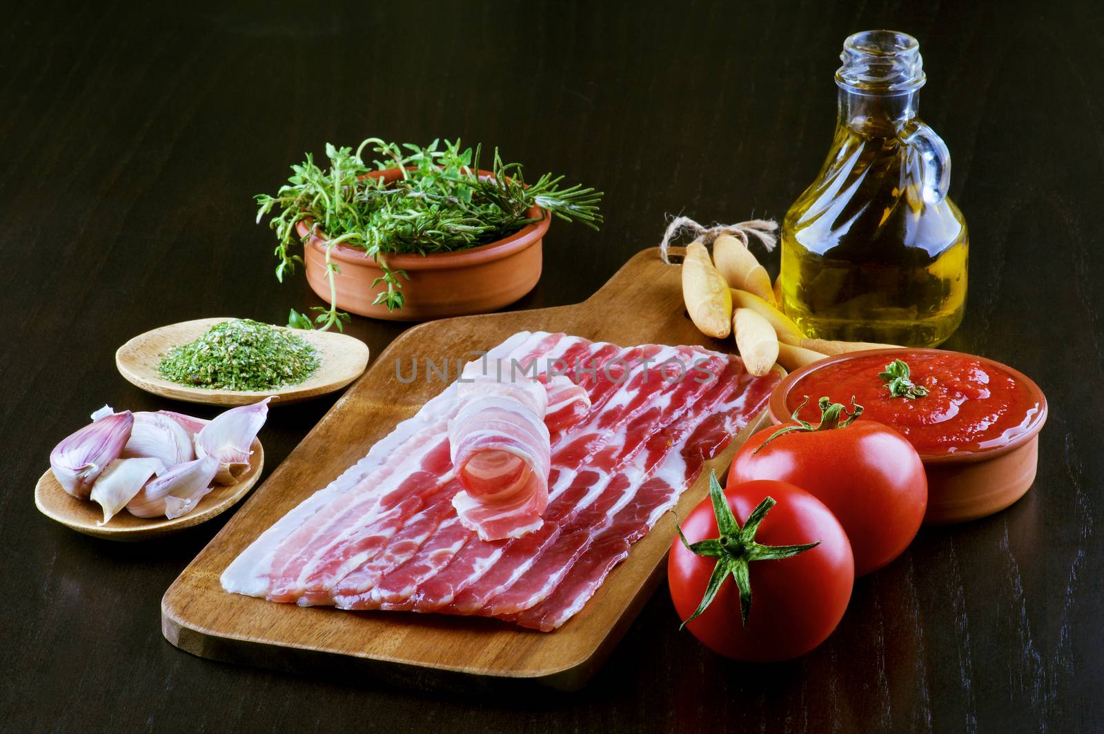 Pancetta on Wooden Cutting Board and Ingredients with Tomatoes, Garlic, Rosemary, Tomato Sauce, Bread Sticks, Spices and Olive Oil closeup on Dark Wooden background