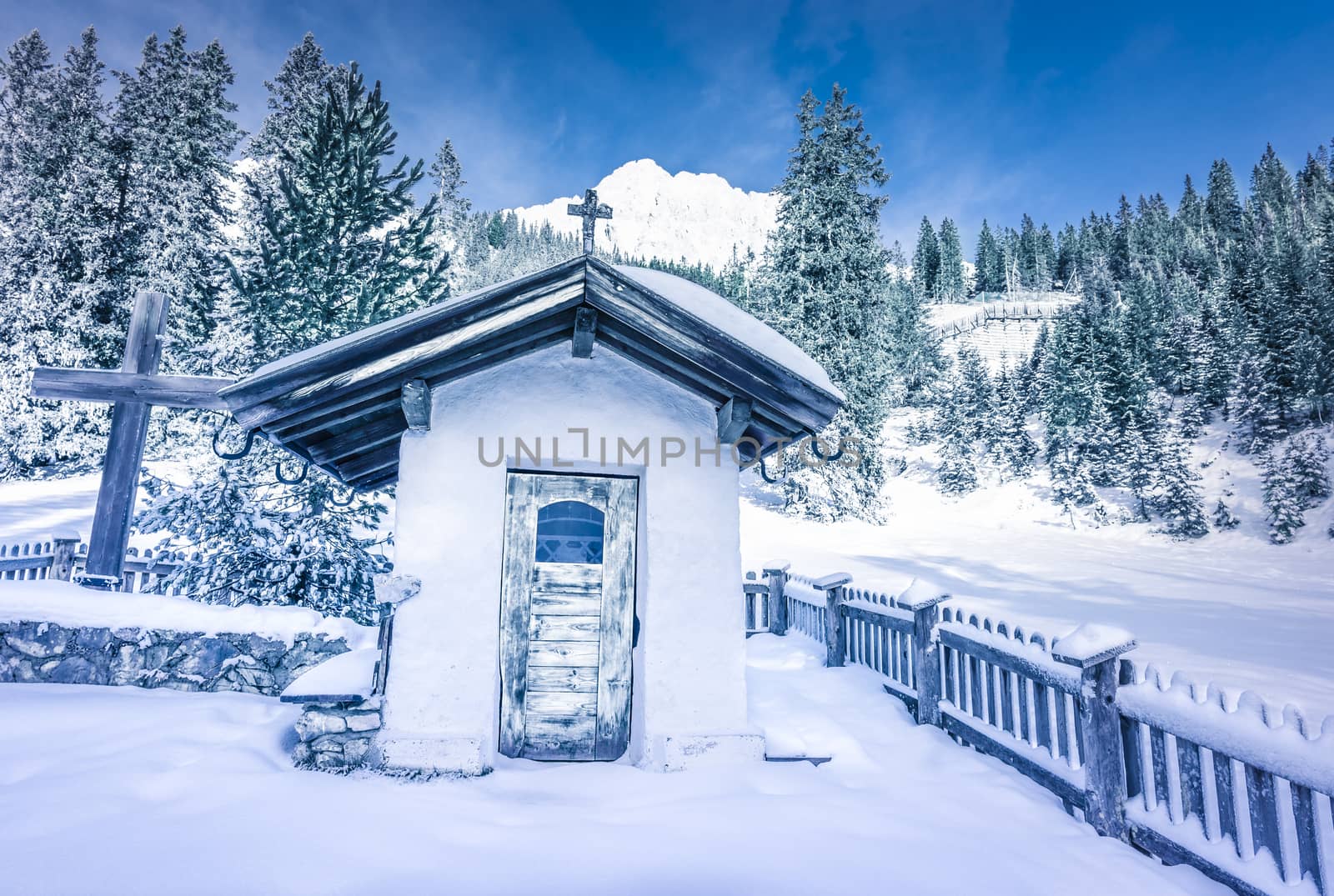 Picturesque snowy scenery in the Austrian Alps with an old chapel  surrounded by an aged wooden fence, a fir forest and mountain peaks covered in snow.