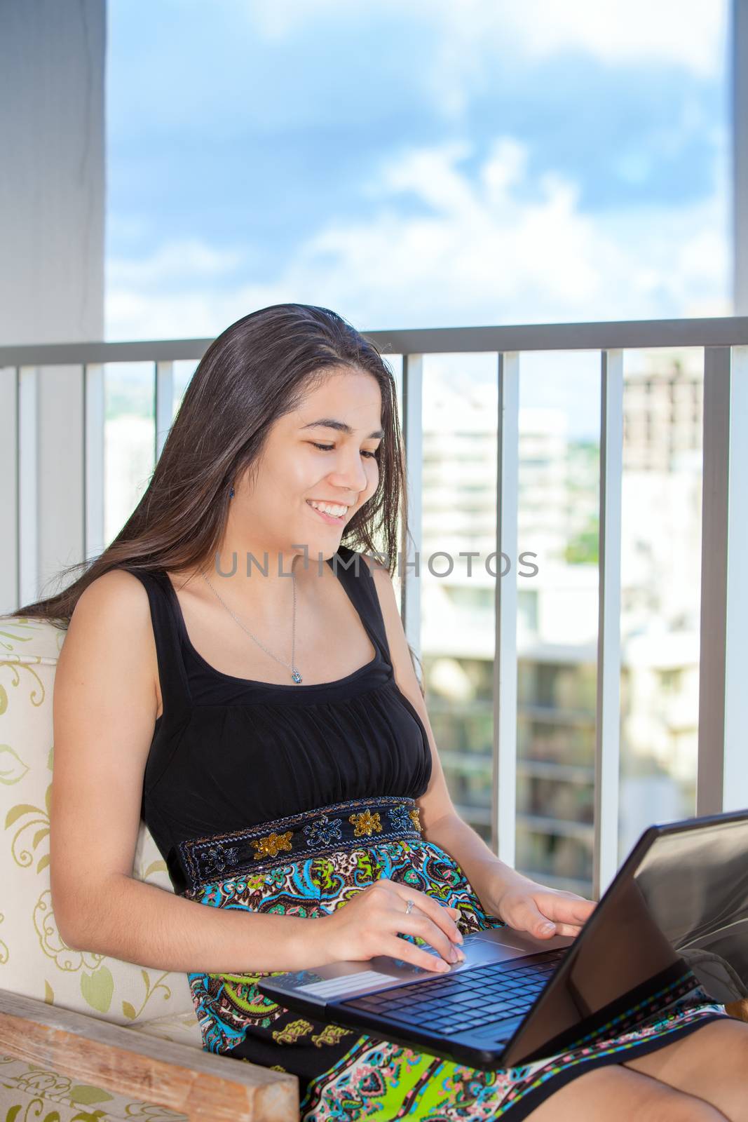 Beautiful teen girl sitting on balcony on highrise building in large city using laptop computer