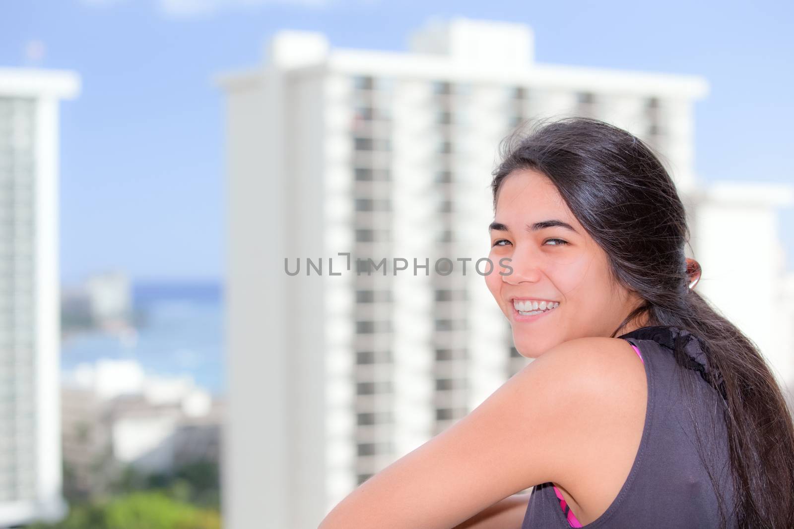 Beautiful biracial teen girl smiling over her shoulder while standing on outdoor balcony with high rise buildings in background of urban city