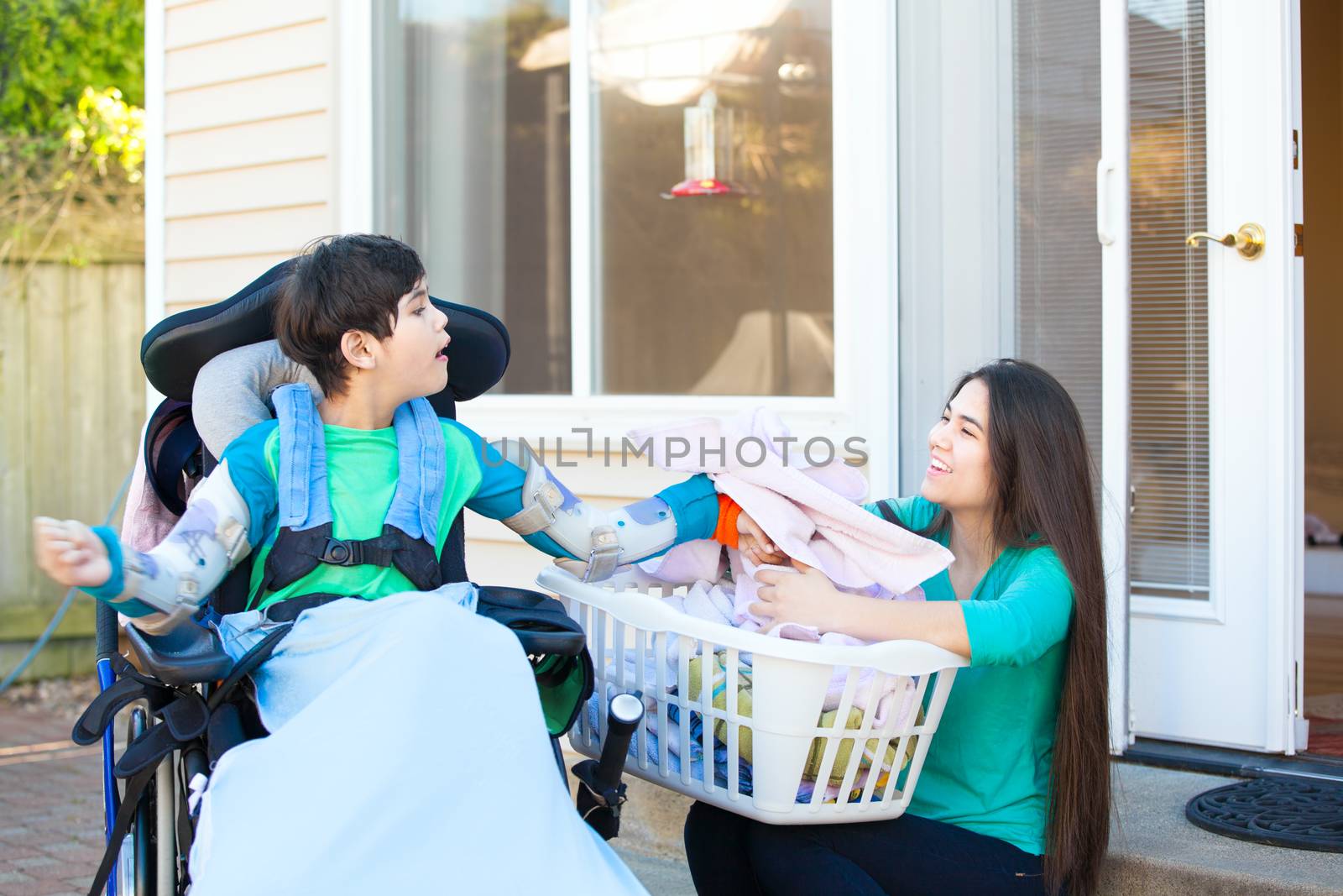 Disabled biracial ten year old boy in wheelchair helping older sister fold laundry on patio outdoors