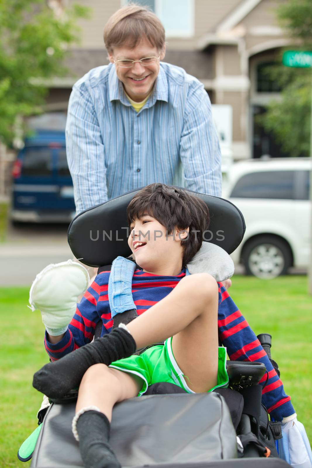 Caucasian father pushing disabled biracial  ten year old son in wheelchair outdoors. Child has cerebral palsy.