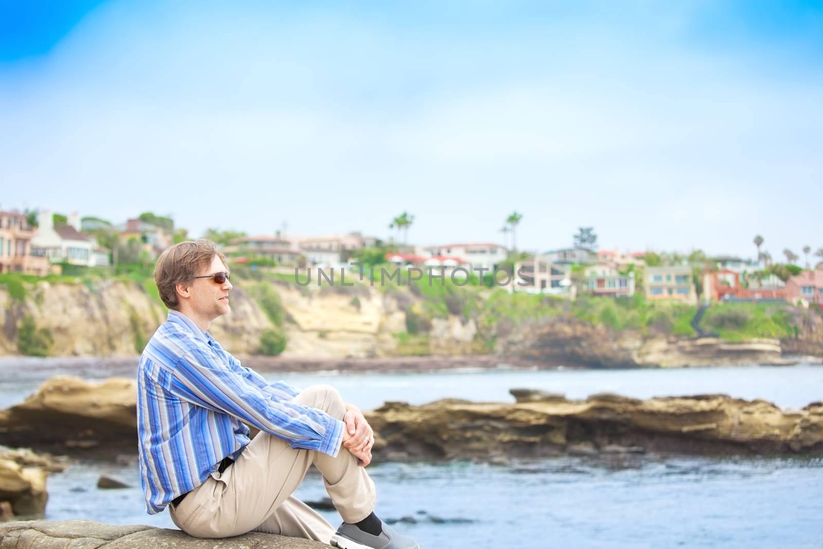 Caucasian man in mid forties sitting  by water's edge with rocky cliff in background, hugging bent knee and looking out over water