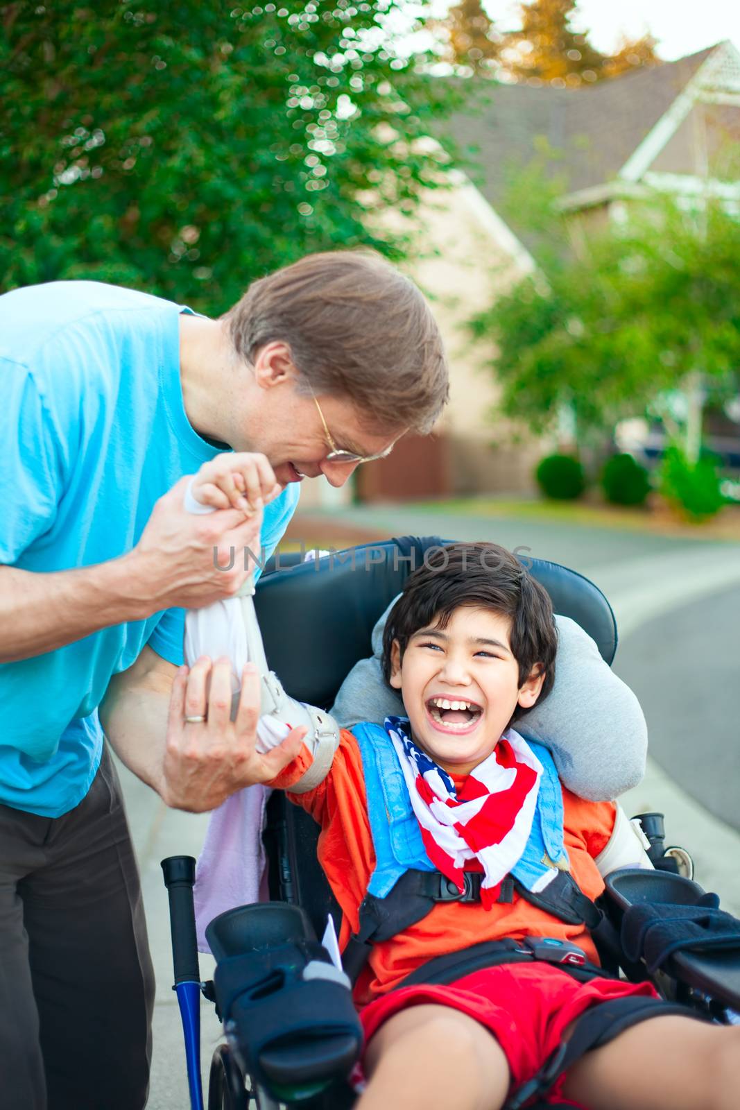 Caucasian father helping disabled ten year old son in wheelchair adjust orthotic arm guards outdoors. Child has cerebral palsy