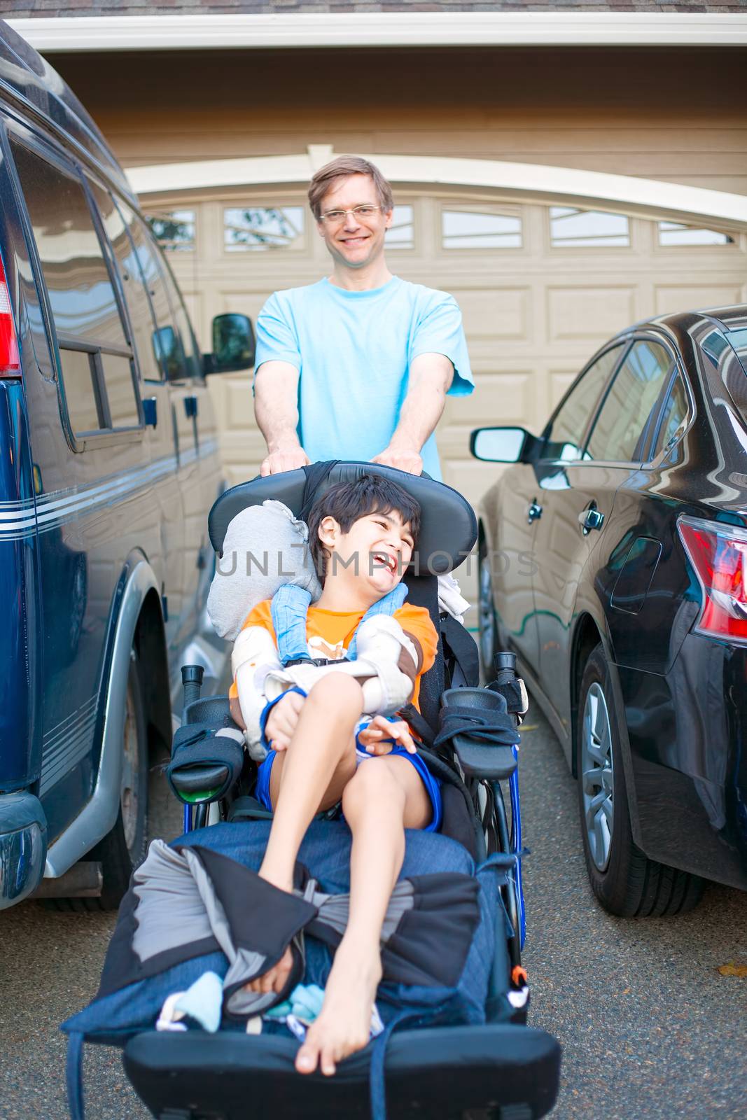 Father pushing disabled ten year old biracial son in wheelchair down driveway past two cars