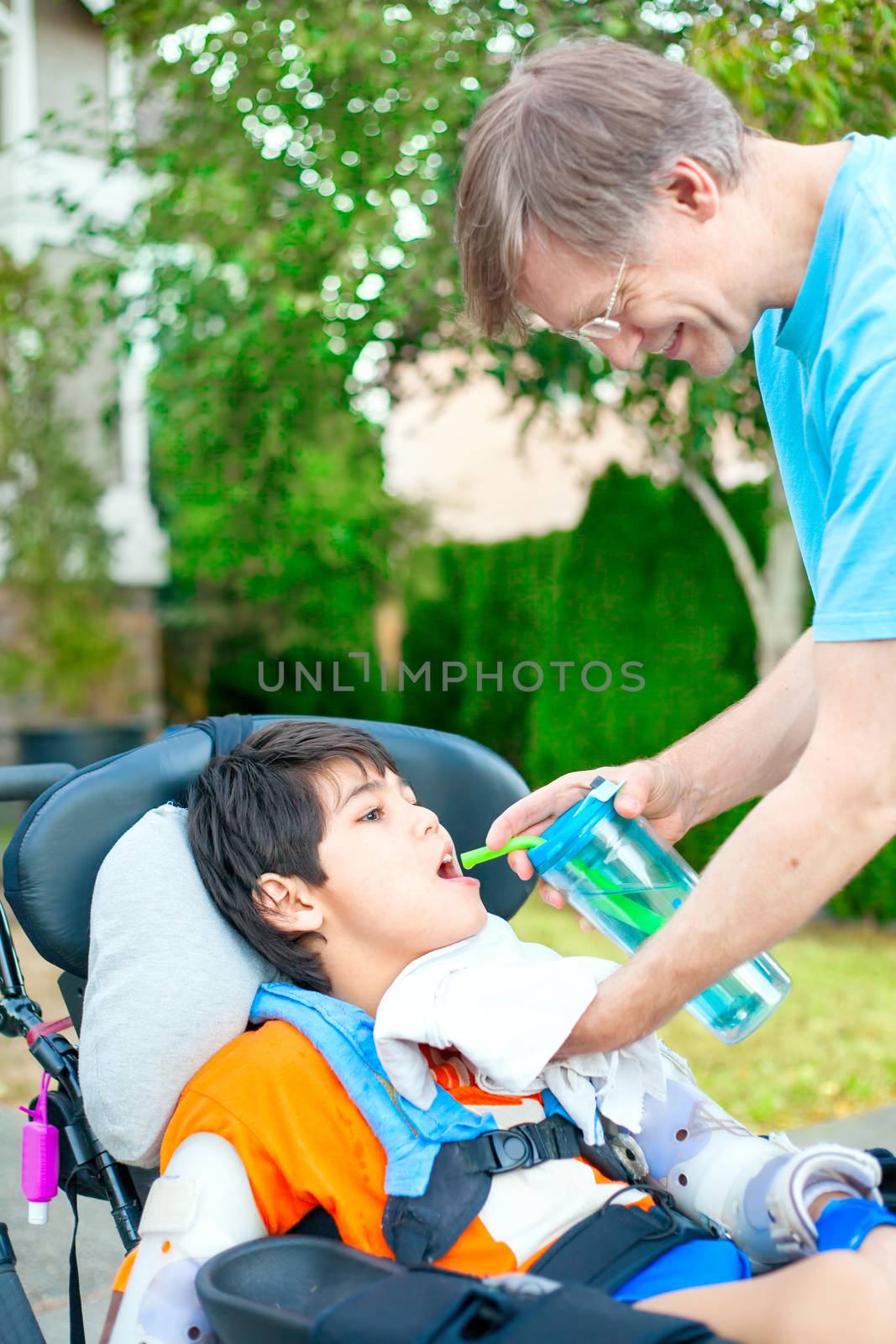 Father holding water cup, helping disabled ten year old son in wheelchair drink from straw