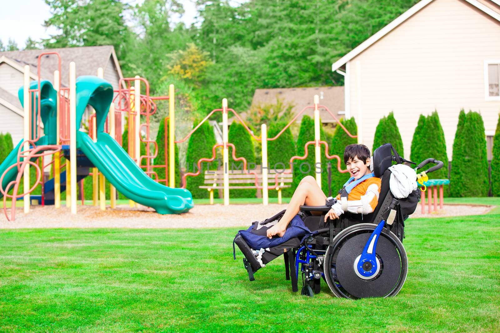 Biracial ten year old disabled boy in wheelchair sitting at a park by a playground, smiling in chair