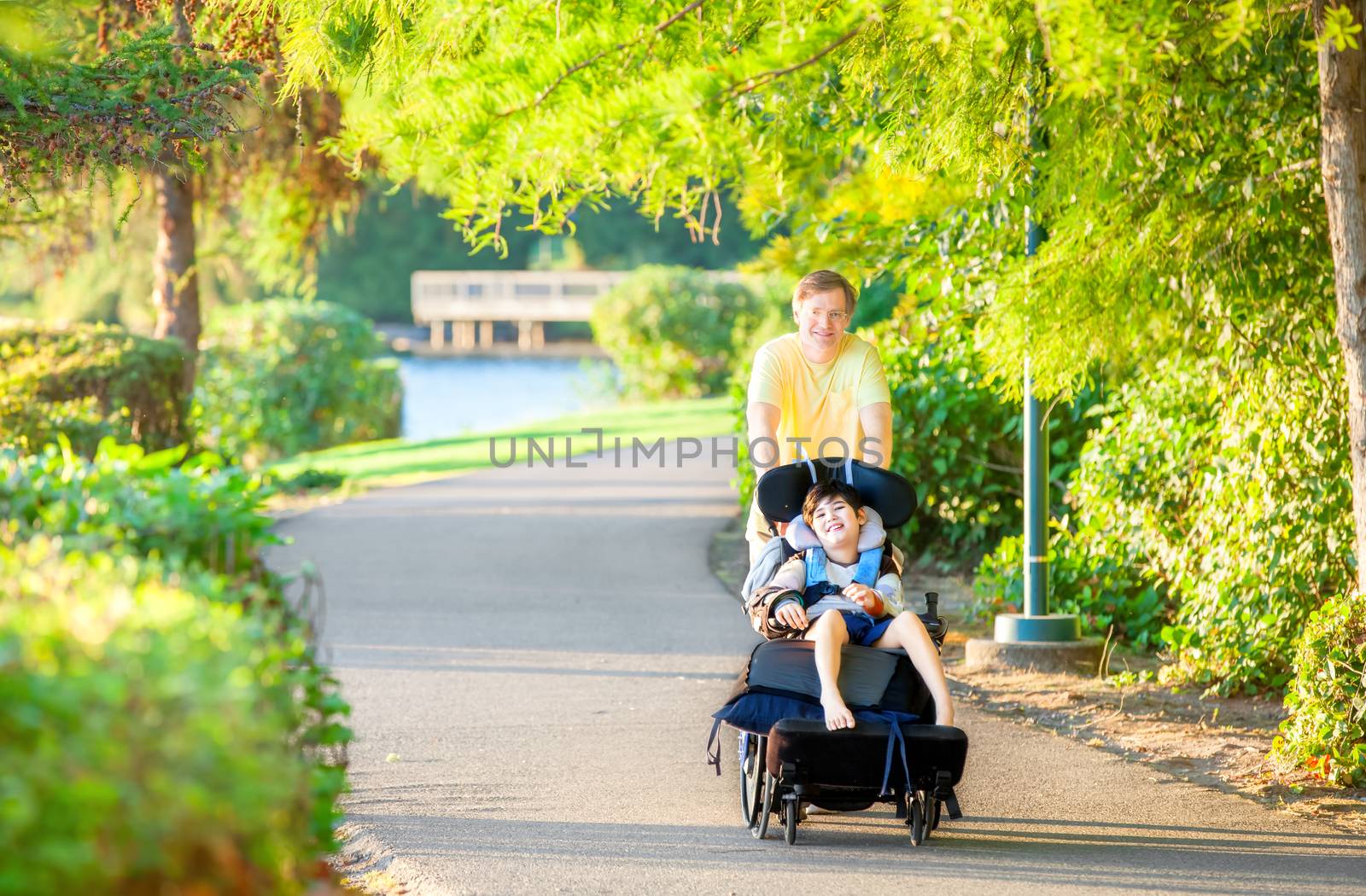 Caucasian father taking walk with ten year old biracial disabled son in wheelchair at park on sunny day