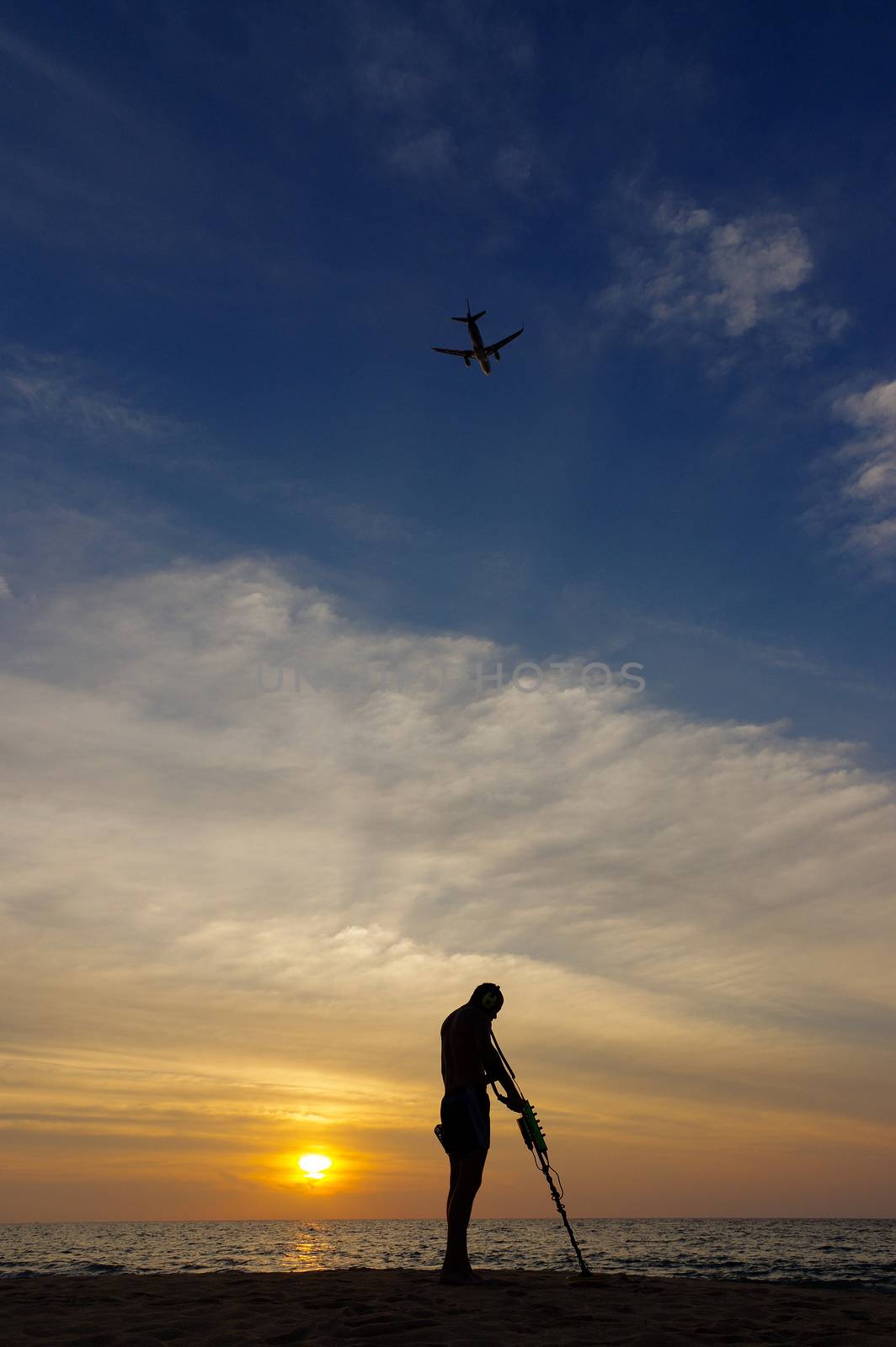 Treasure hunter with Metal detector on the sunset the beach,  a plan in the sky. by evolutionnow