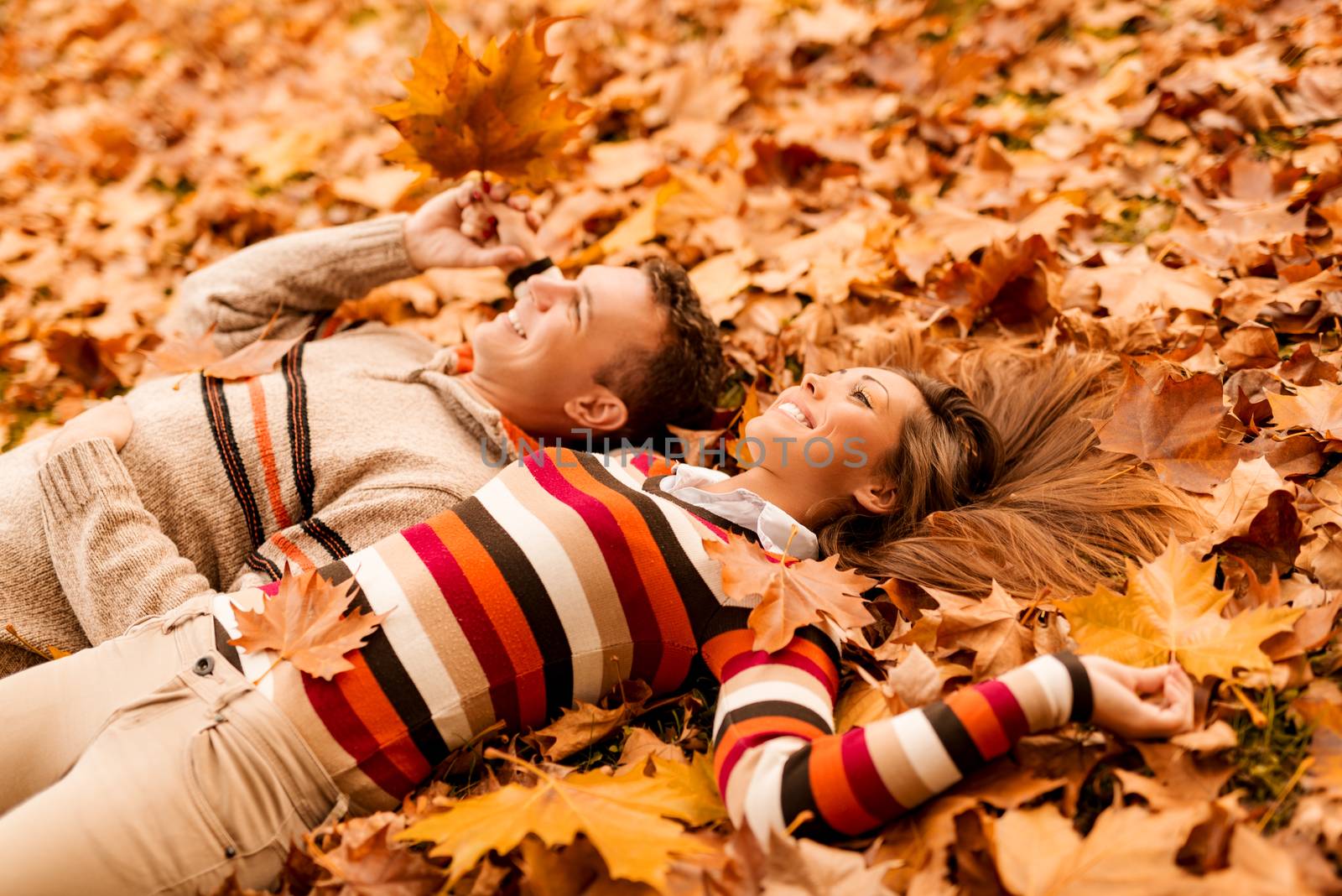 Beautiful smiling couple enjoying in sunny forest in autumn colors. They are lying on the falls leaves.