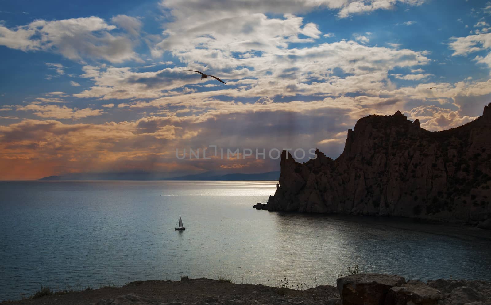 Evening landscape with sunset and clouds over a calm sea, cliffs, seagulls and a sailboat, Crimea, Novy Svet.