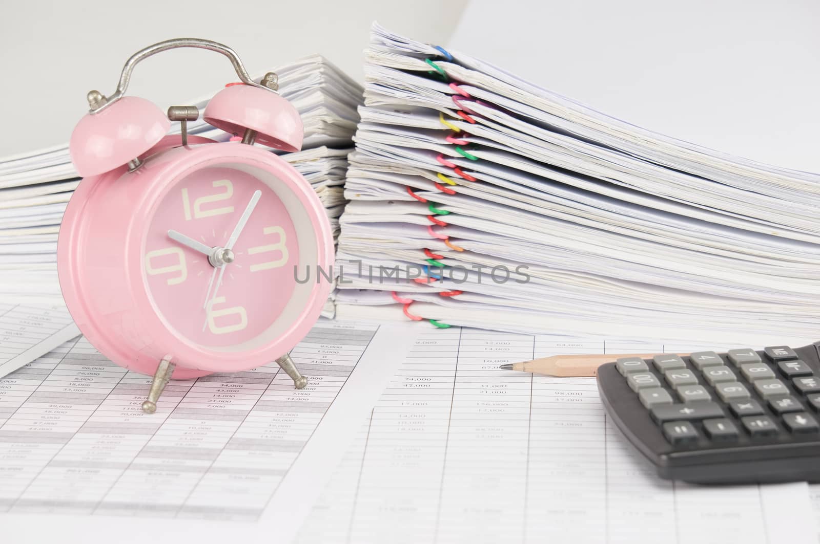 Brown pencil and old pink alarm clock on finance account have blur calculator and pile overload paperwork of report and receipt with colorful paperclip as background.