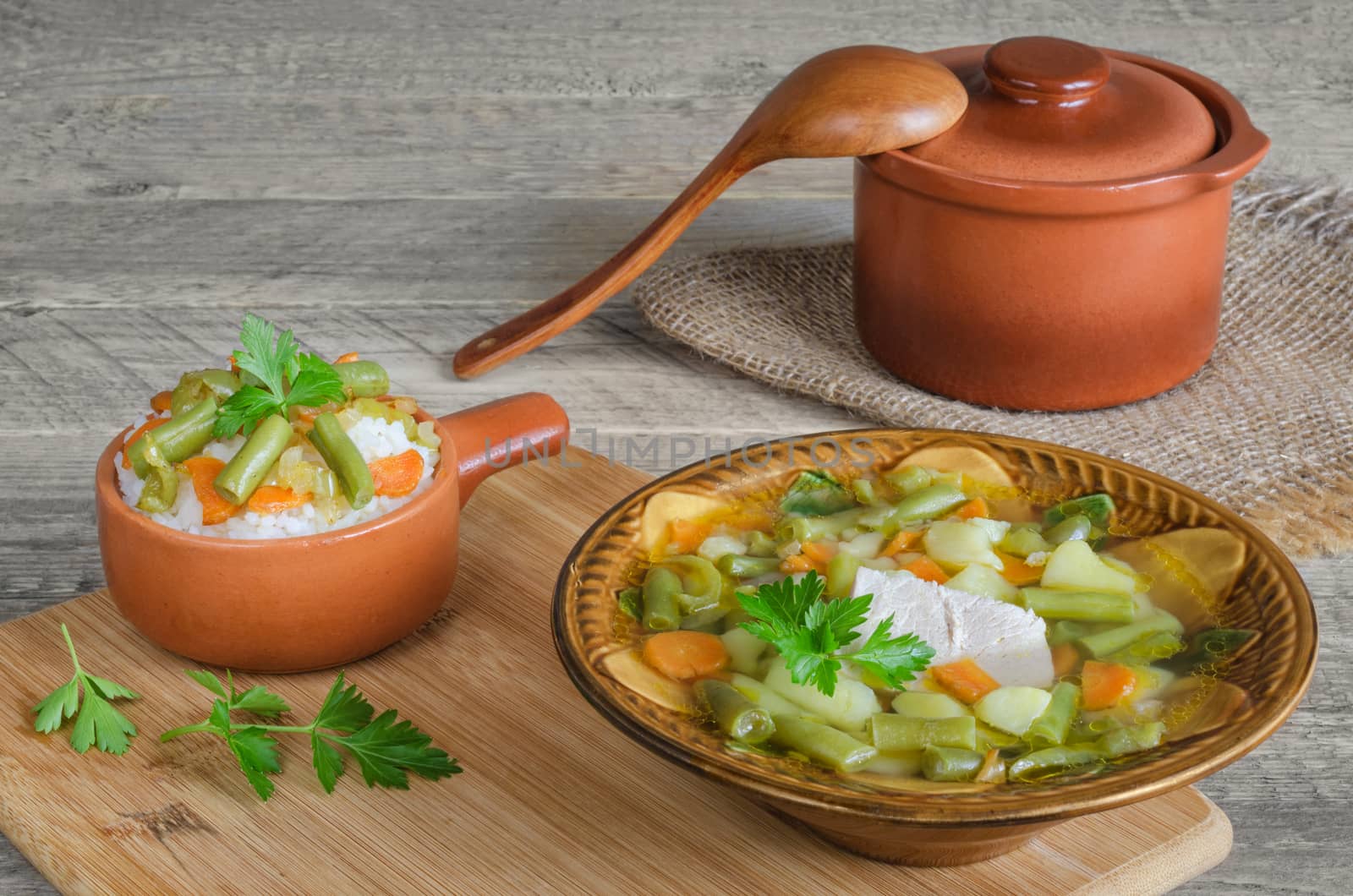 Soup of pork with vegetables and rice, ceramic pot  spoon, on wooden background. by Gaina