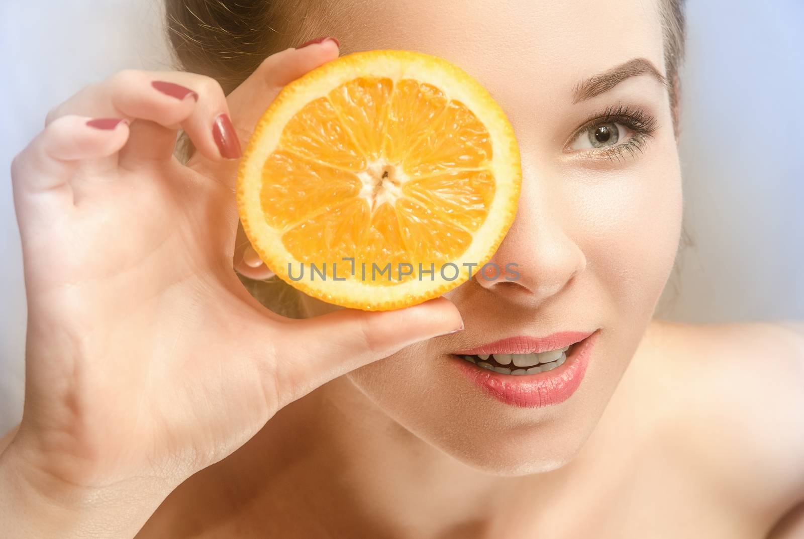 Young beautiful woman with perfect skin holding a slice of orange in front of her eyes and smiles looking ahead. Half-turn face. Gray background