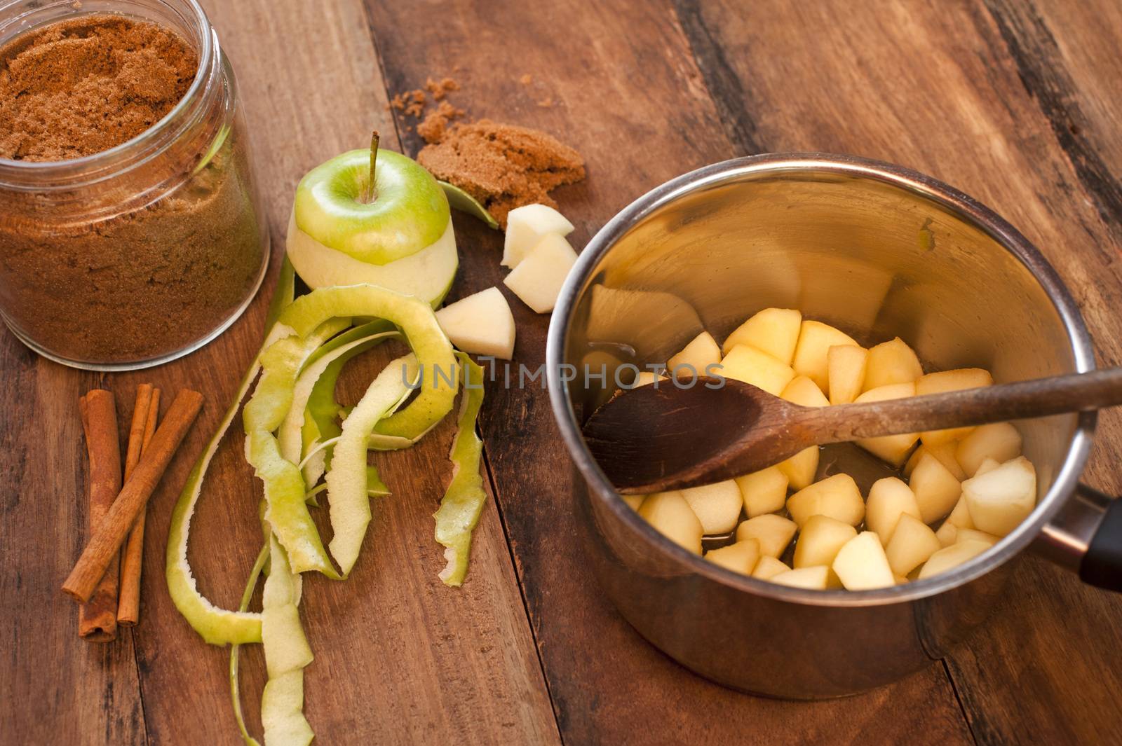 Preparing homemade apple and cinnamon sauce with fresh peeled and sliced green apples in a pot alongside stick cinnamon and ground spice on a wood table