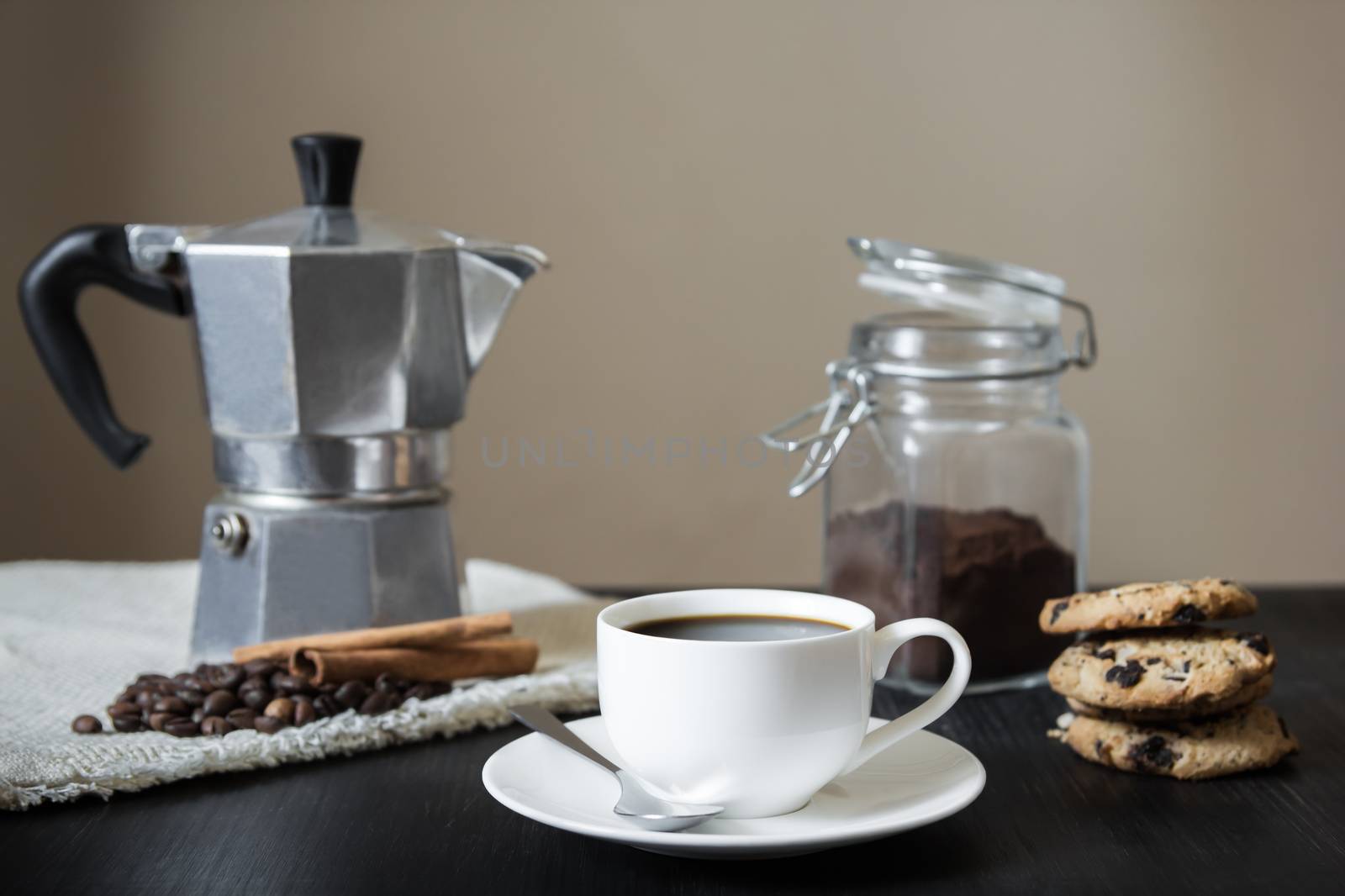 Front view of cup of black coffee, italian coffee pot on flax table-napkin and chocolate chip cookies on black vintage wood table indoors in natural light